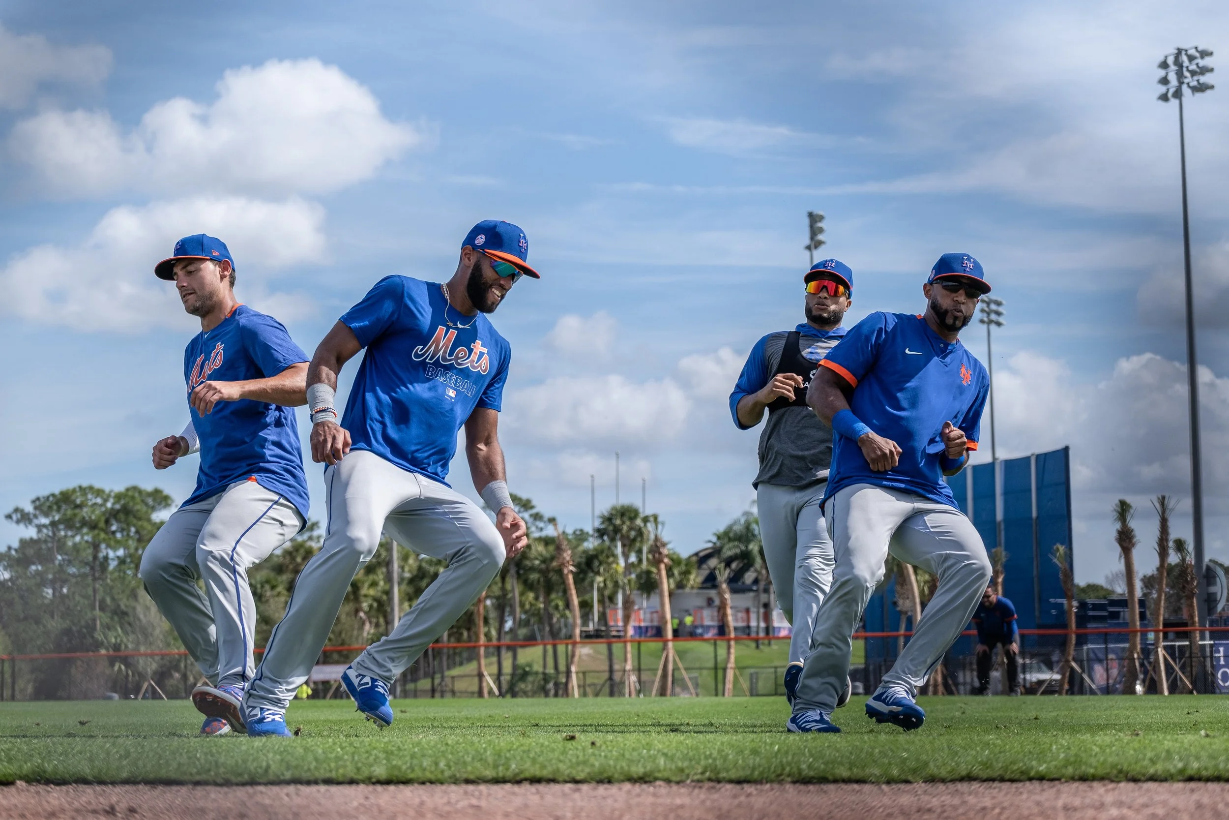 Four baseball players in blue jerseys and white pants practicing on a field under a partly cloudy sky.