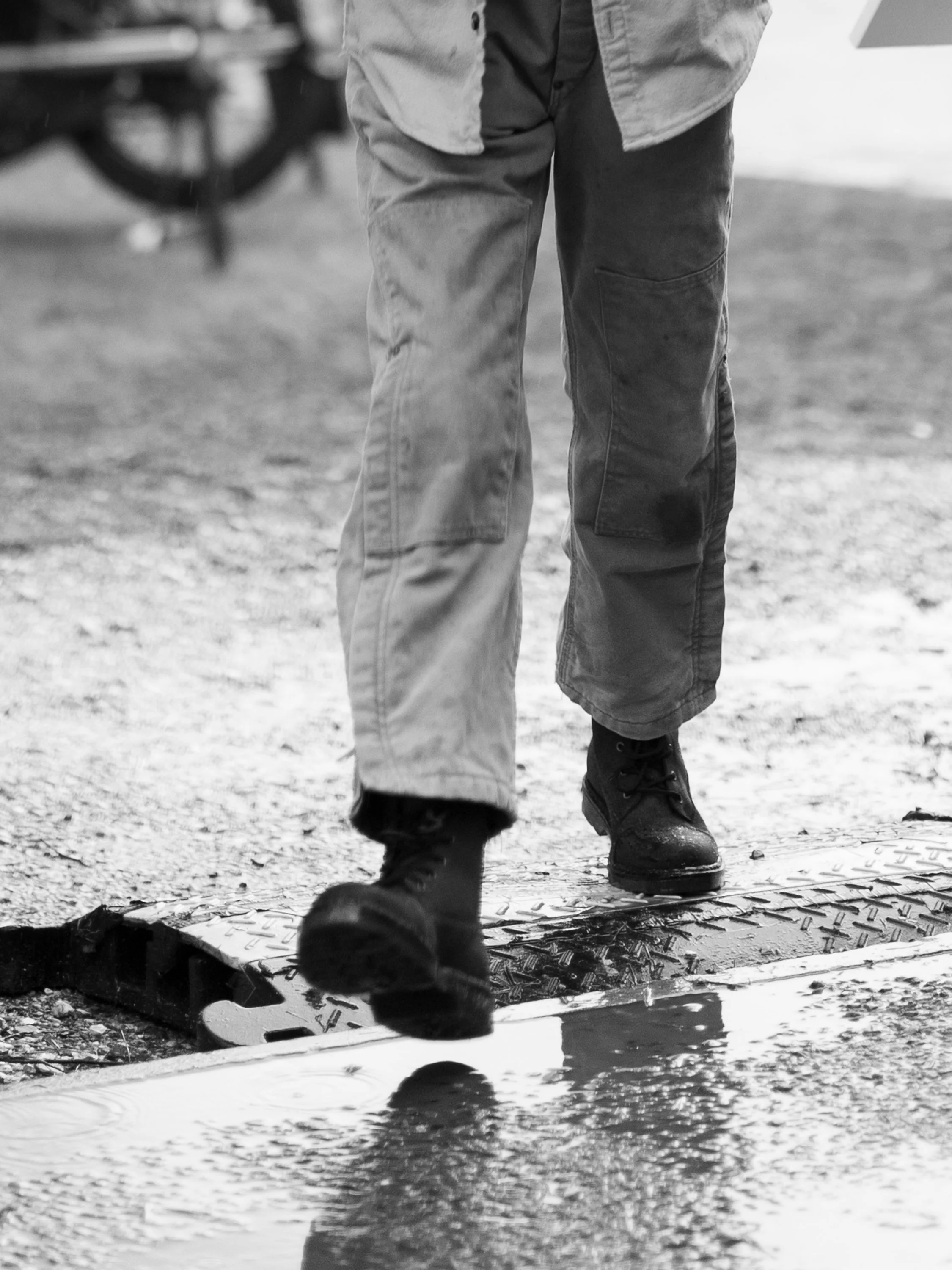 Close-up of a person walking over a water drain cover on a textured ground, wearing dark tactical boots, baggy pants, and a button-up shirt, with a blurred outdoor background.