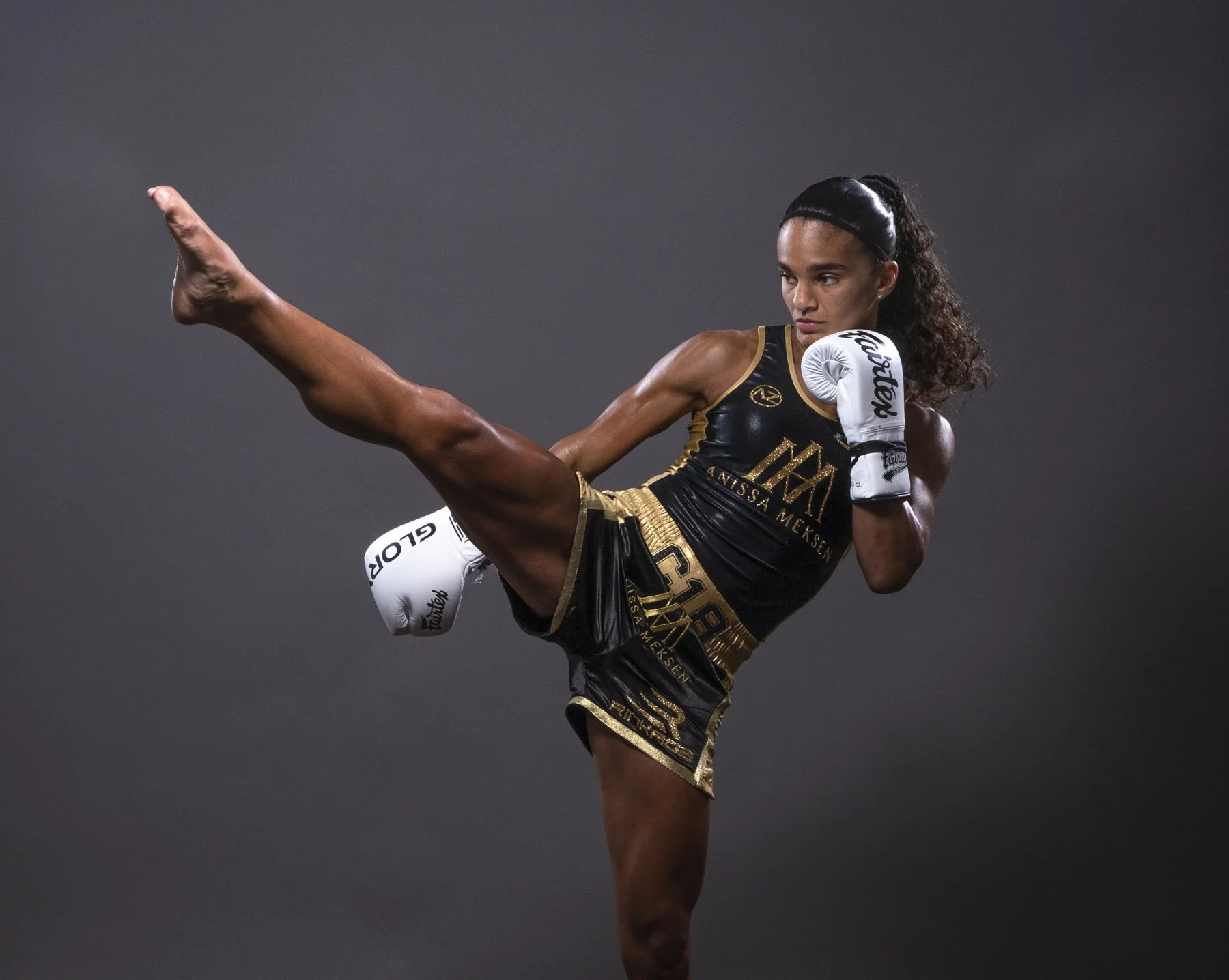 Female martial artist in black and gold uniform executing high kick with gloves, dark background.