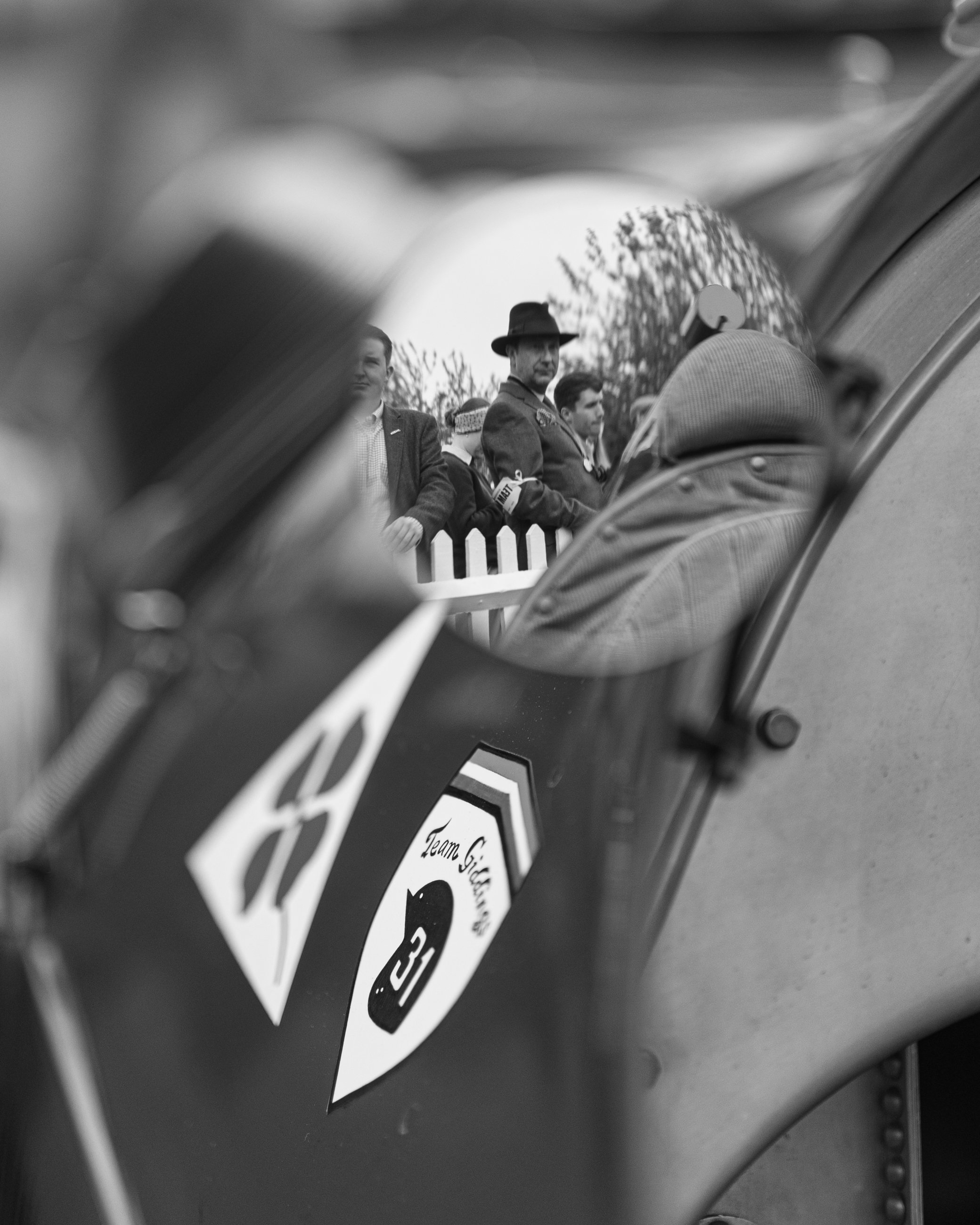 A group of people dressed in vintage military and civilian attire, standing outdoors near a white picket fence with trees in the background, as seen through a circular mirror reflecting part of the scene. Goodwood Revival