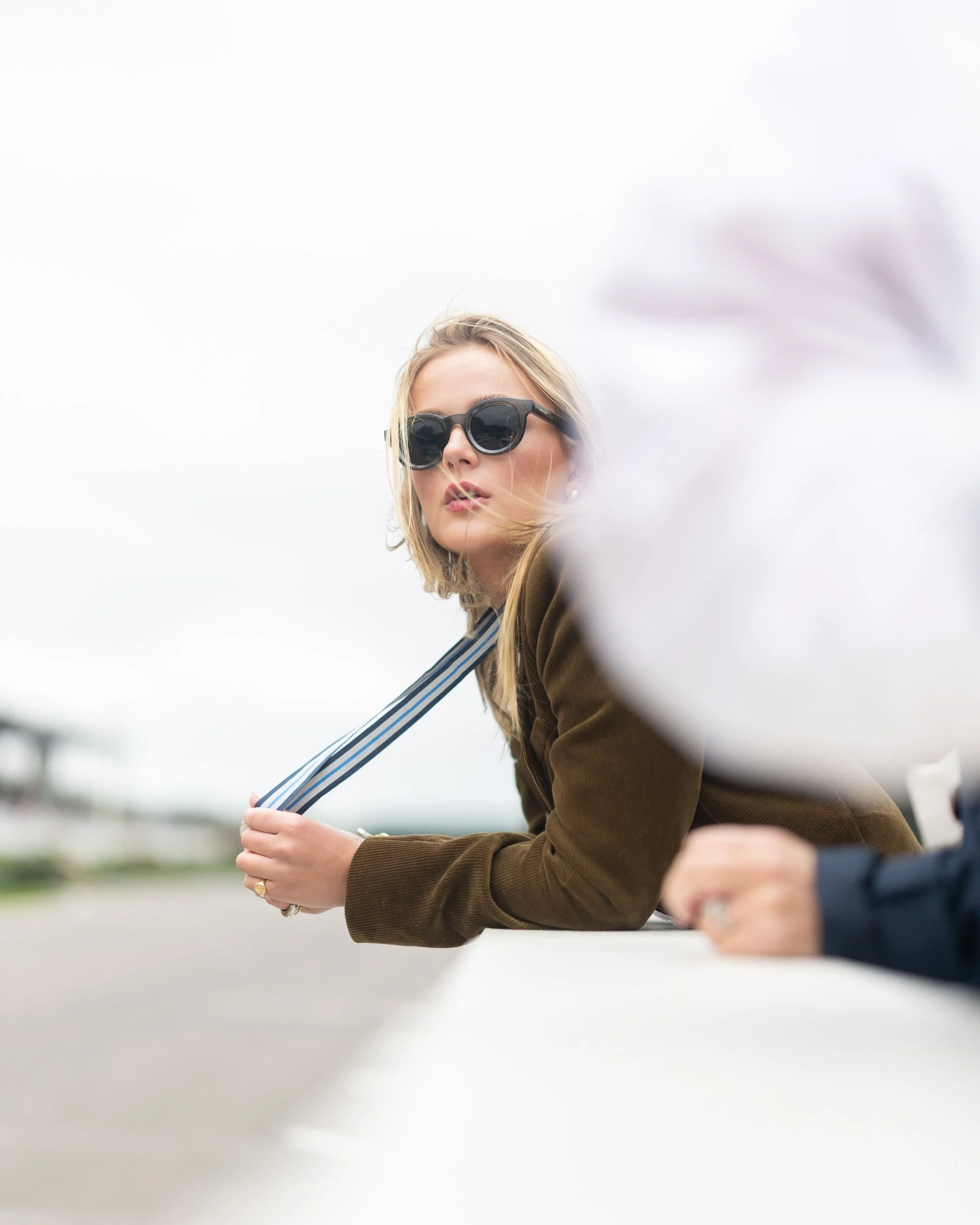 A woman with blonde hair wearing dark sunglasses and a brown jacket, leaning on a surface outdoors with an overcast sky in the background.