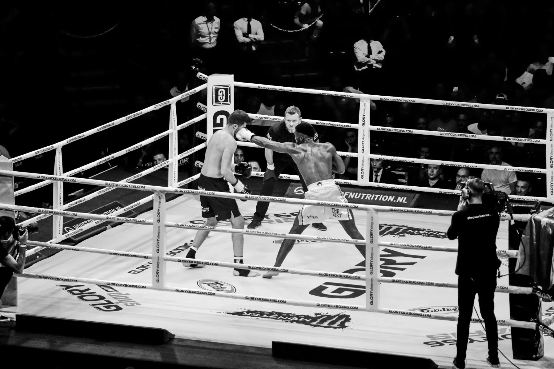 Two male fighters are engaged in a boxing match inside a ring with three factors, with a referee observing closely. One fighter is throwing a punch at the other, who is wearing boxing gloves and shorts, while several spectators and photographers are 