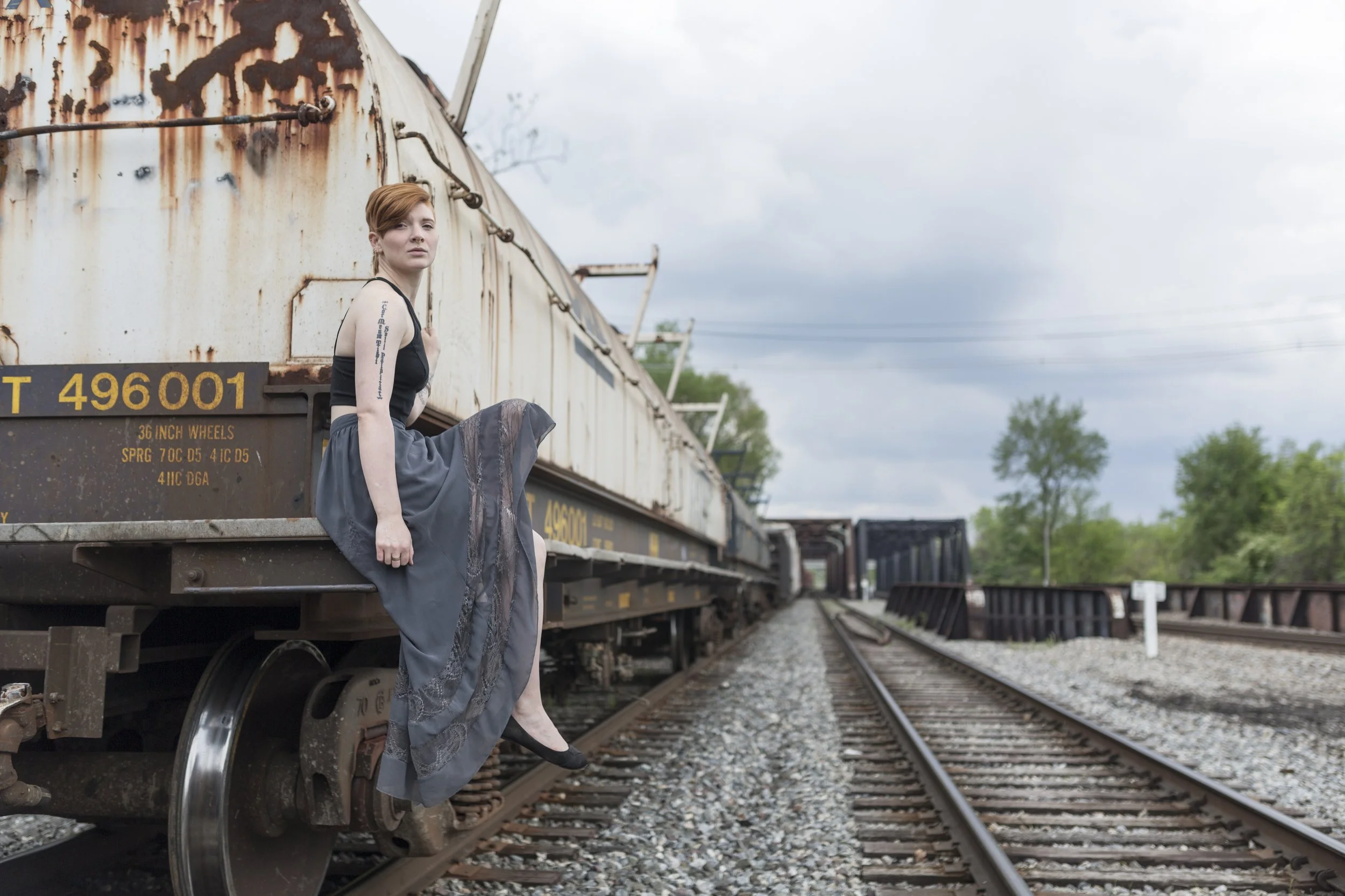 A young woman with red hair and a tattoo on her arm, wearing a black top and gray skirt, sitting on the edge of a rusty train car on a railway track, with her leg hanging off. The background shows cloudy skies and trees.