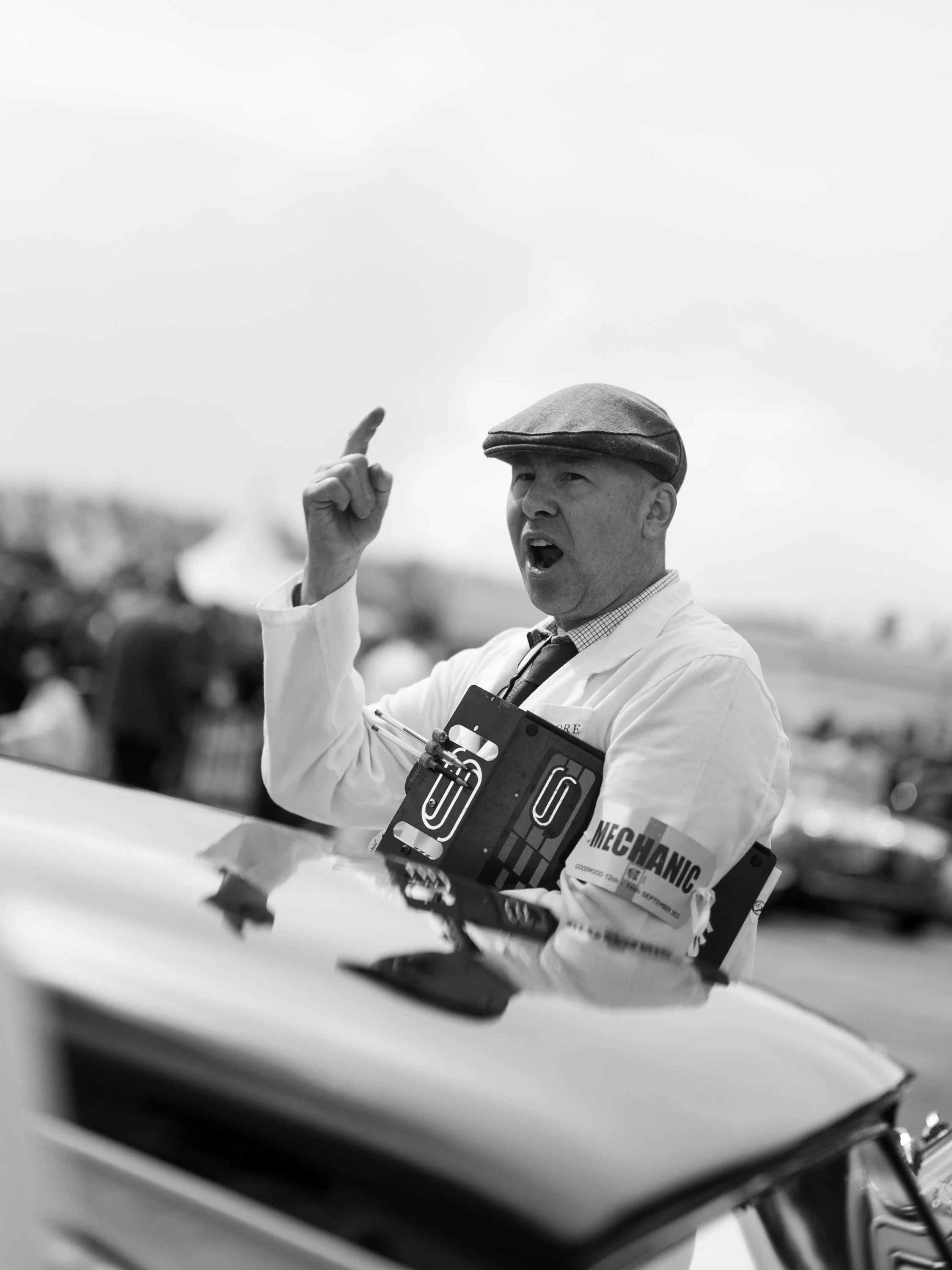A man in a flat cap and white blazer gesturing with his right hand while speaking, with a clear sky in the background.