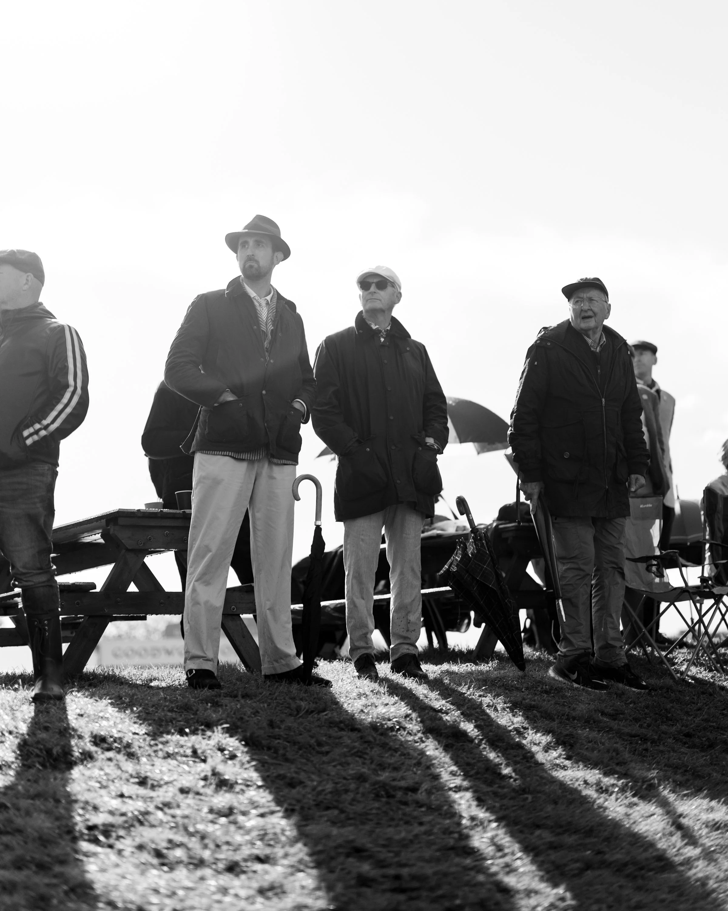 Four men standing outdoors on a grassy field, facing forward. They are wearing jackets and hats, and one has sunglasses, with shadows cast on the ground in front of them.