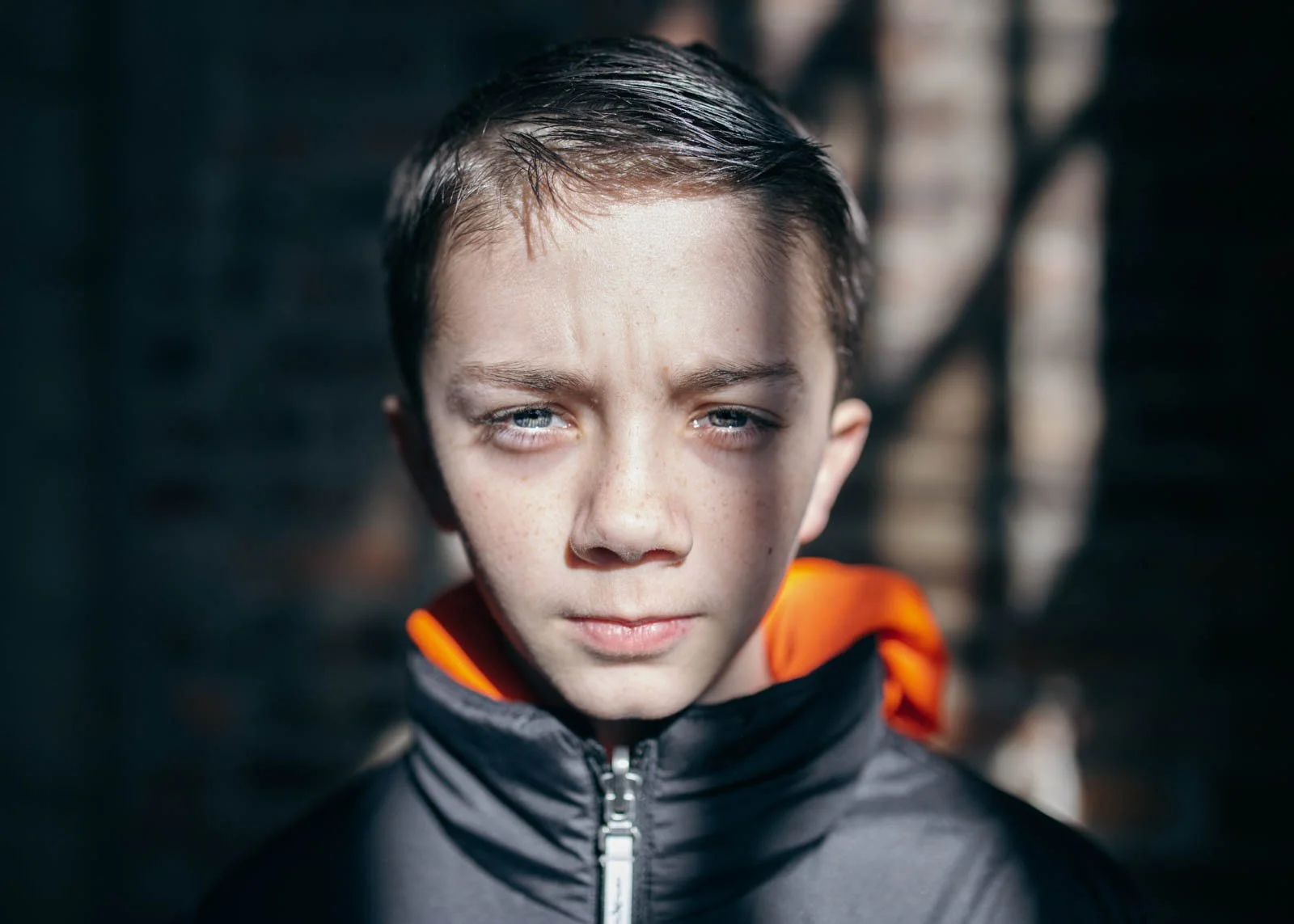 Close-up of a young boy with wet brown hair, wearing a black jacket with an orange lining, standing outdoors with a blurred background.