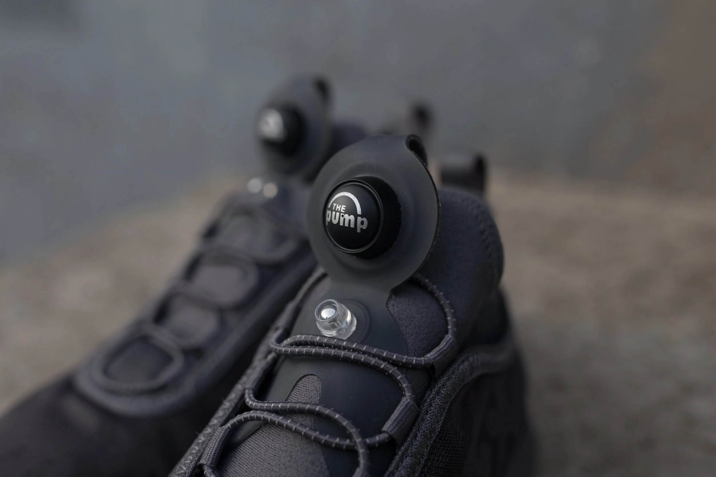 Close-up of a black tactical glove holding a firearm with a button that says 'The Pump' on the grip, the background is blurred. Reebok.