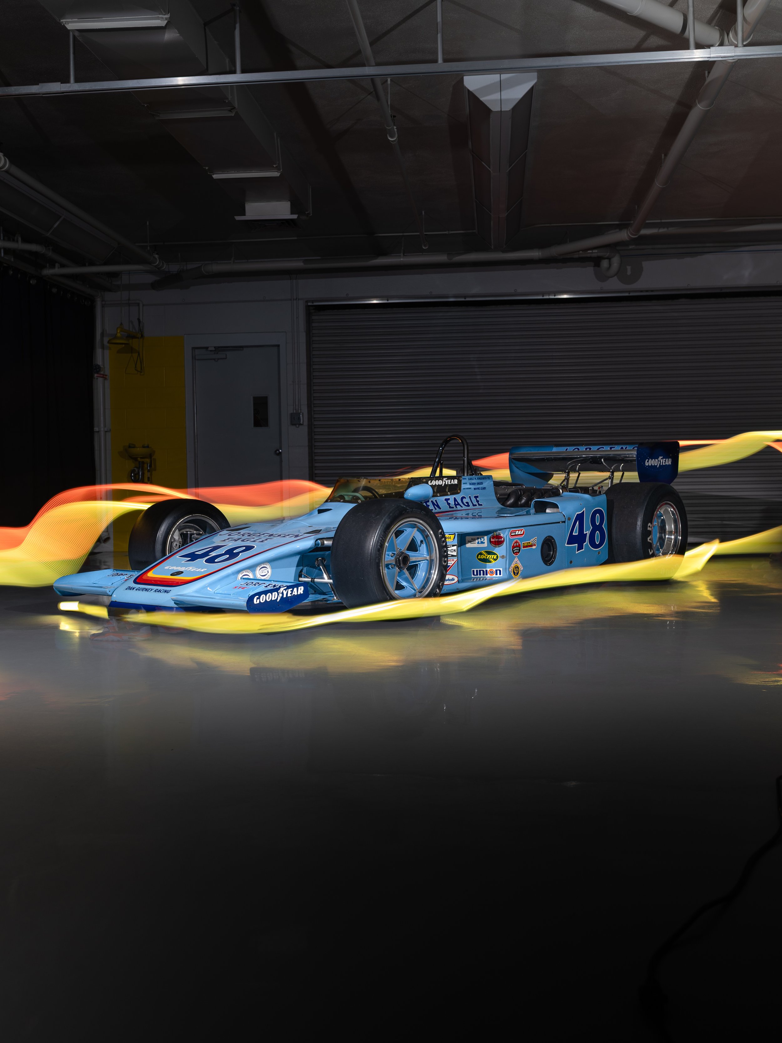 Classic race car with the number 48, decorated with various sponsor logos, in a garage or underground parking area with industrial ceiling pipes, partially lit with dramatic lighting effects.