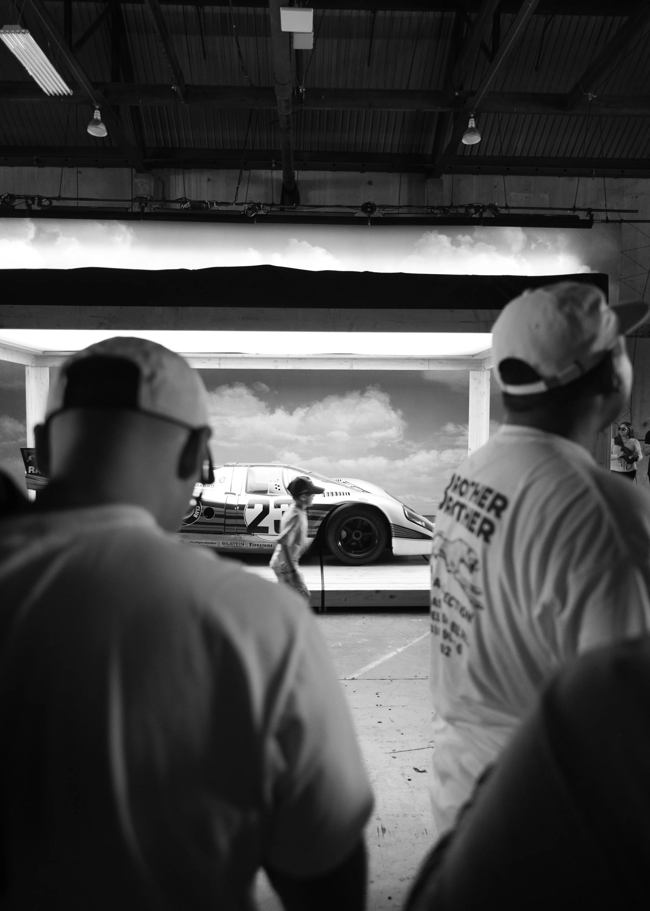 Black and white photo of race car team members in garage overlooking race car on track, with open garage door showing sky and clouds outside.