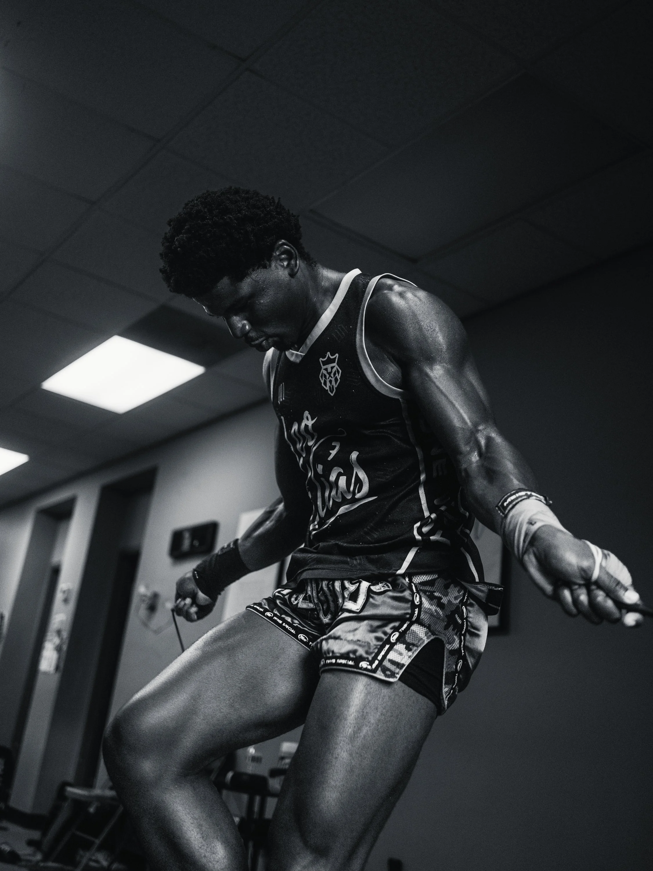 A male athlete in a gym, wearing a sports jersey and shorts, leaning forward with sweat on his skin, holding gym equipment, with a low ceiling and fluorescent lights above.