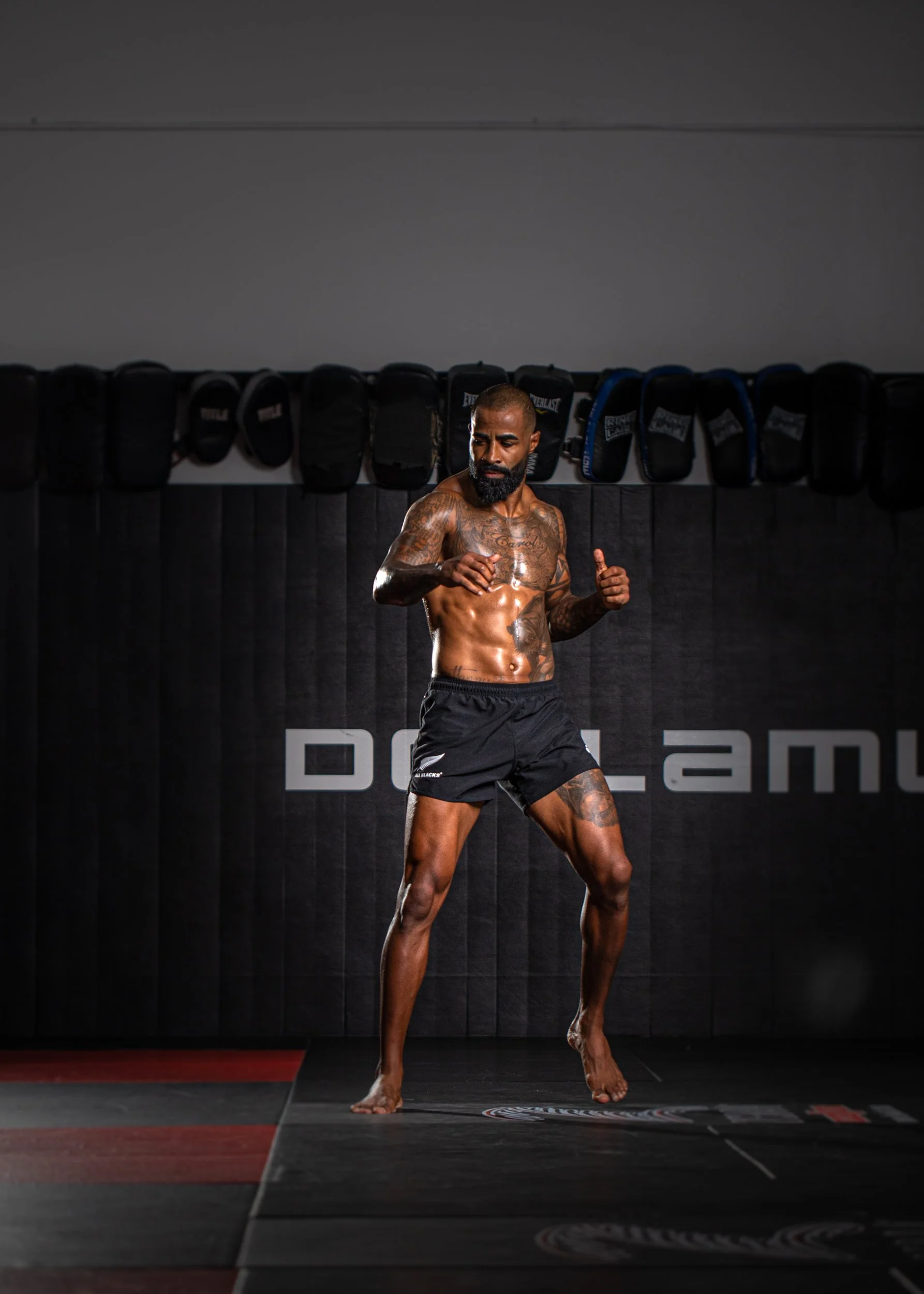 A muscular man with tattoos on his chest and arms, wearing black shorts, stands shirtless on a gym mat in a training facility. There are black and blue punching bags hanging on the wall behind him, and the environment has a dark, intense atmosphere. 