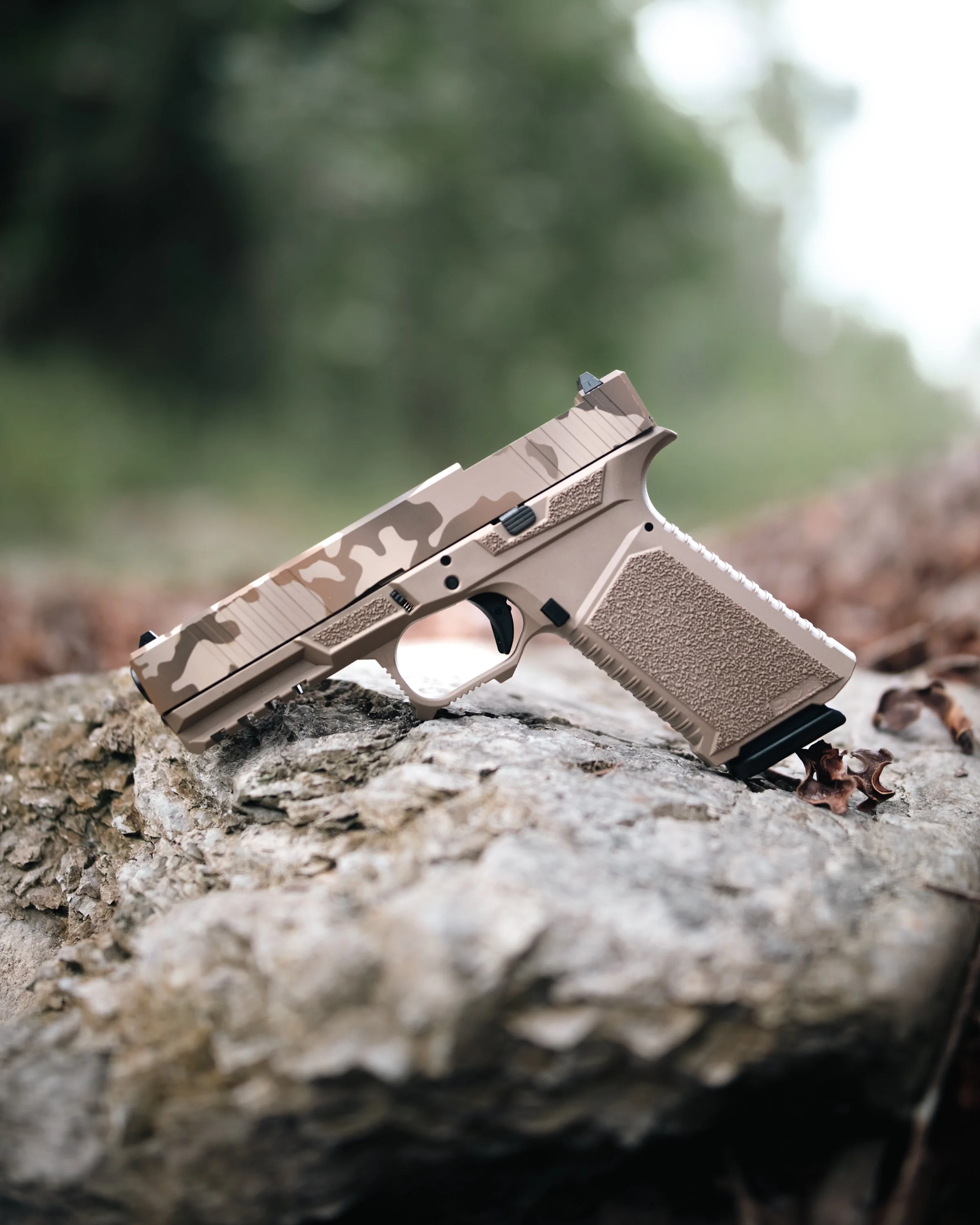 Close-up of a tan and black handgun resting on a rough, textured tree trunk outdoors, with a blurred green background. Photographed for SCT Manufacturing.
