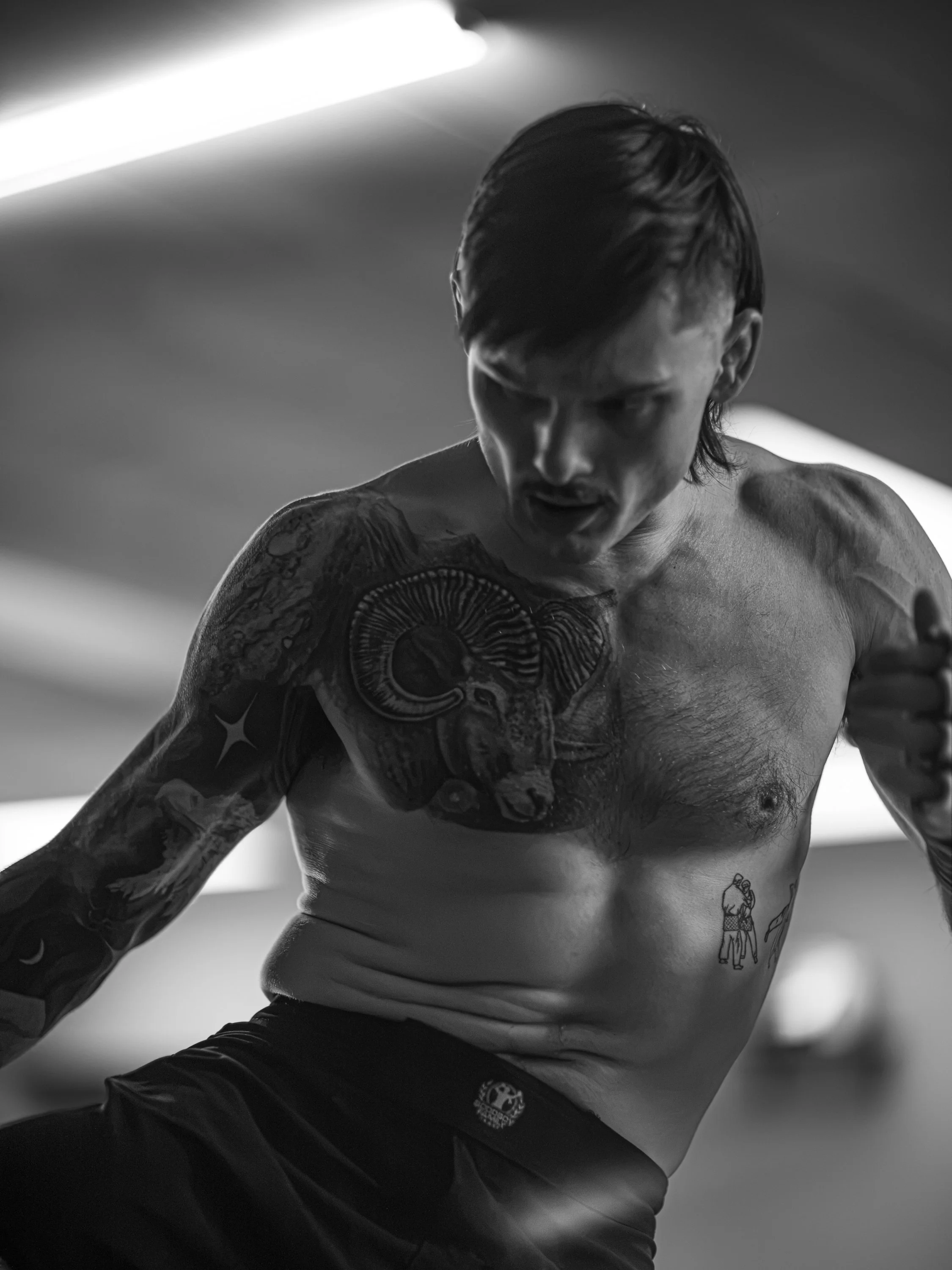 Black and white photo of a shirtless man with tattoos on his arms and chest, looking down with an intense expression, in a gym or studio setting.