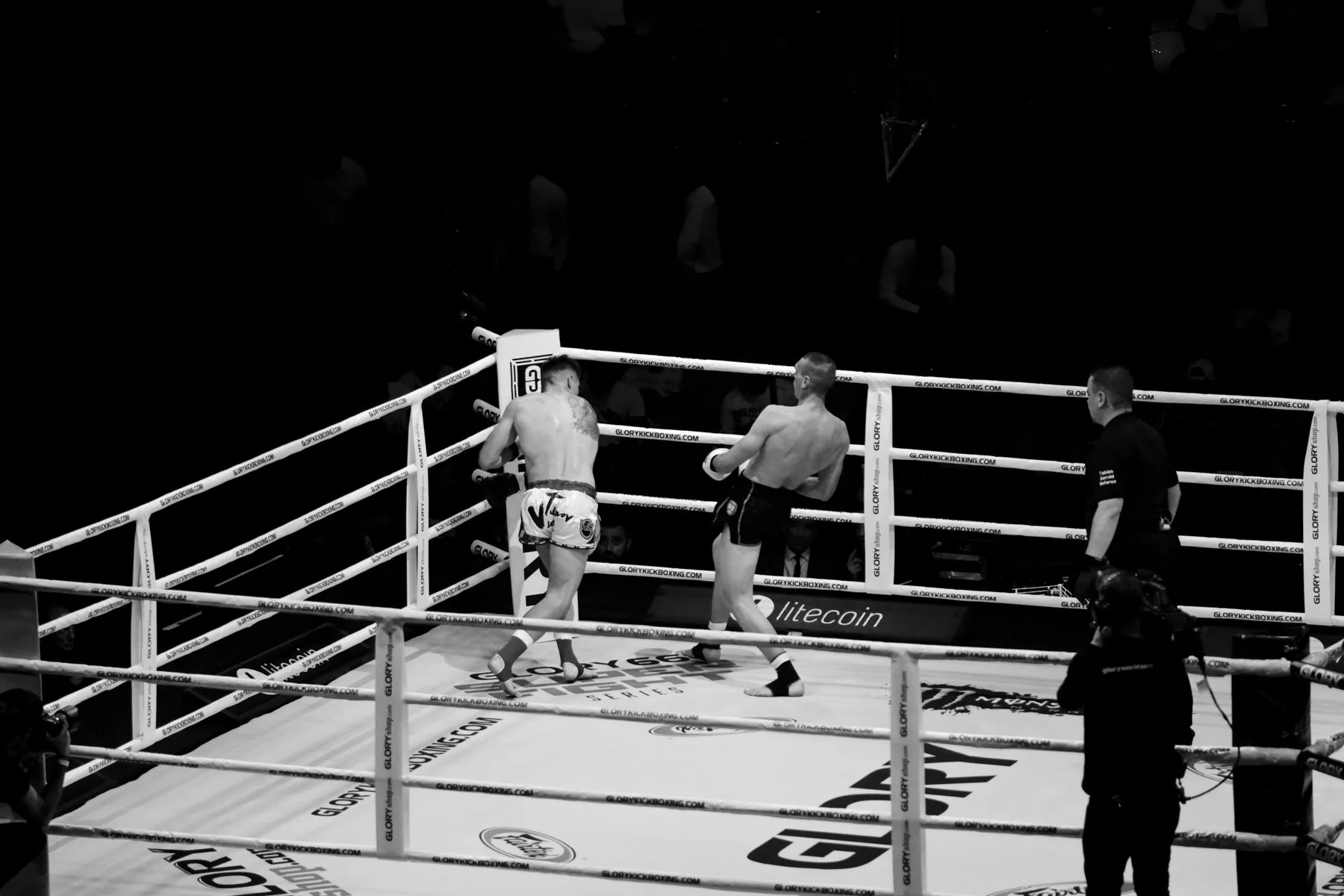 Two fighters in a kickboxing ring, one with tattoos and the other in black shorts, facing off as a referee looks on. The ring has advertising banners, including Litecoin and Glory Kickboxing.