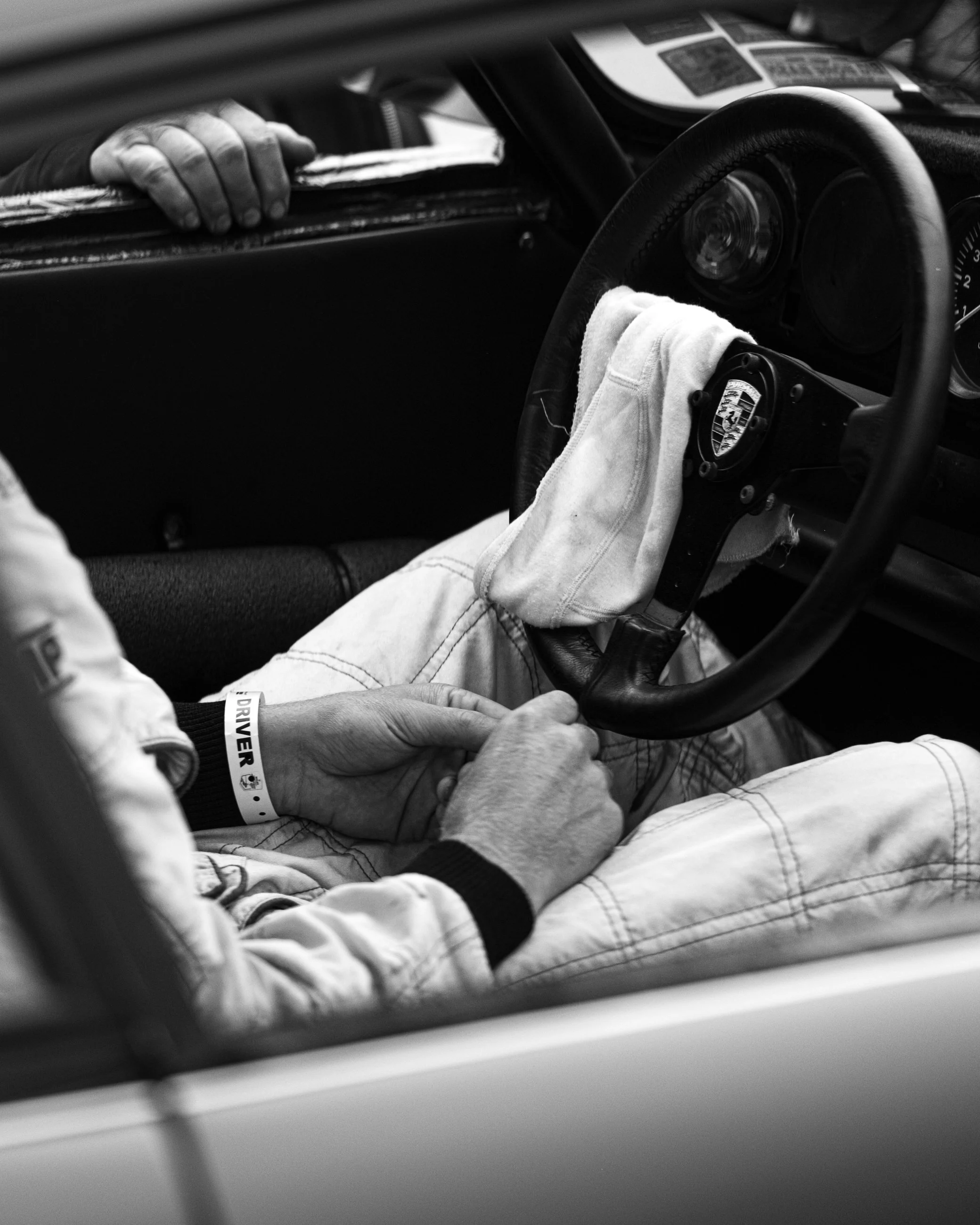 Close-up of racing car interior with a hand resting on a stack of papers on the dashboard, steering wheel with Porsche emblem, and a towel draped over the gear shifter.