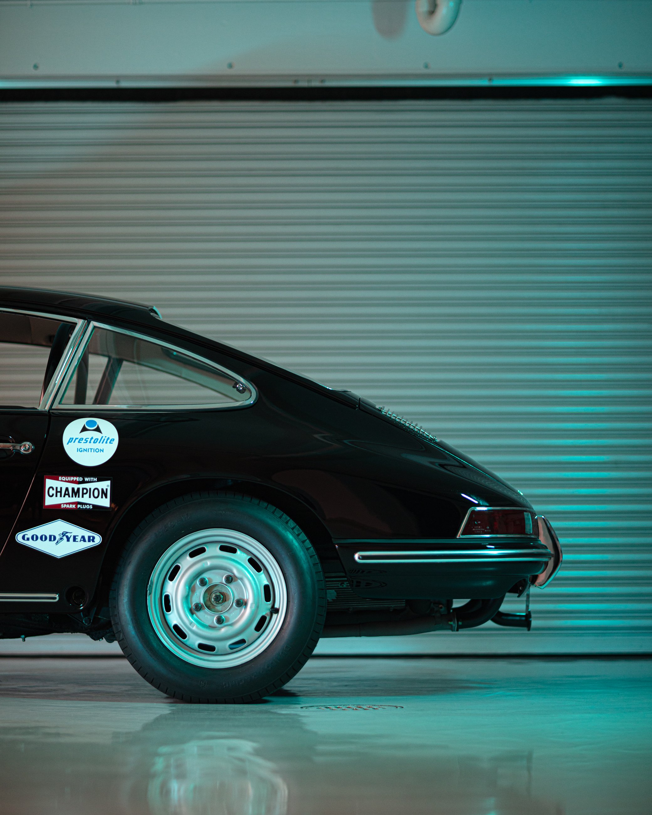 A partial view of a black classic car with stickers on its side, parked on a photo studio floor with a metallic, striped background.