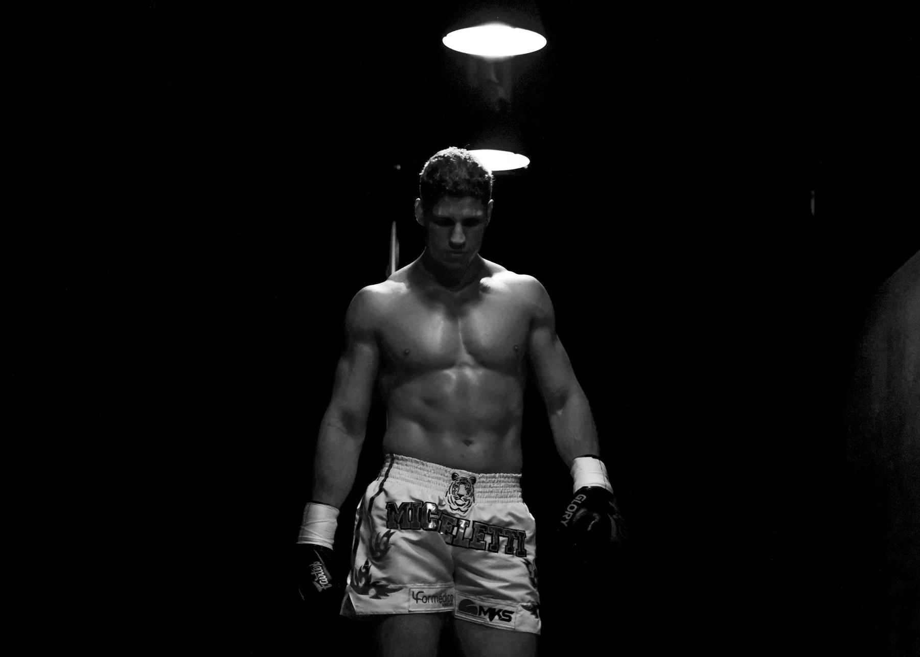 Black and white photo of a shirtless male boxer standing with his head slightly bowed, wearing gloves and shorts with various logos, under overhead lights in a dark setting.