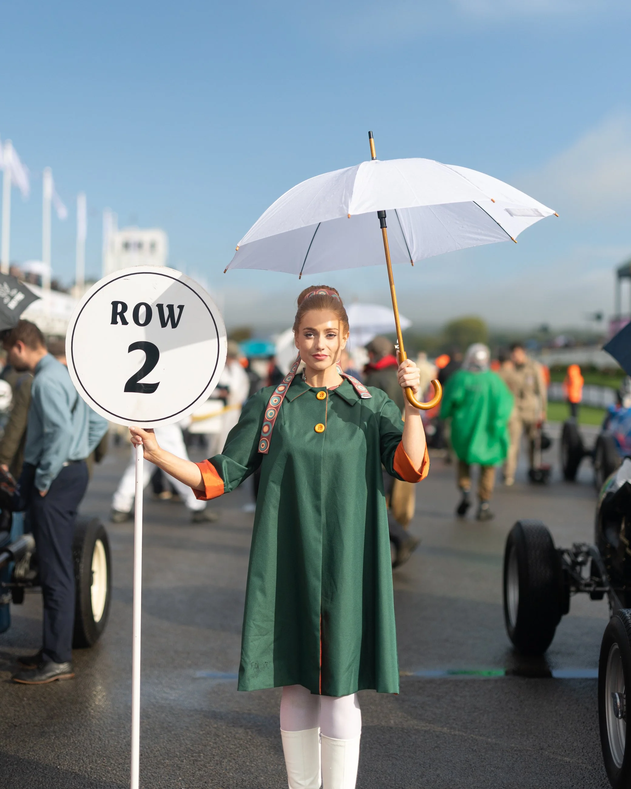 Woman standing on street holding a white sign that reads "Row 2" and a white umbrella, dressed in a green dress with orange accents and white thigh-high socks, with a crowd and blue sky in the background.