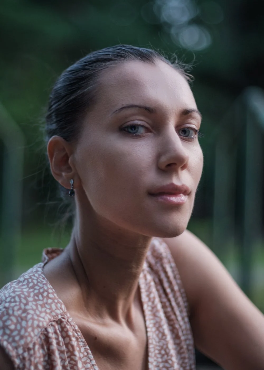 Close-up of a woman with blue eyes, dark hair slicked back, and earrings, outdoors with a blurred green background.