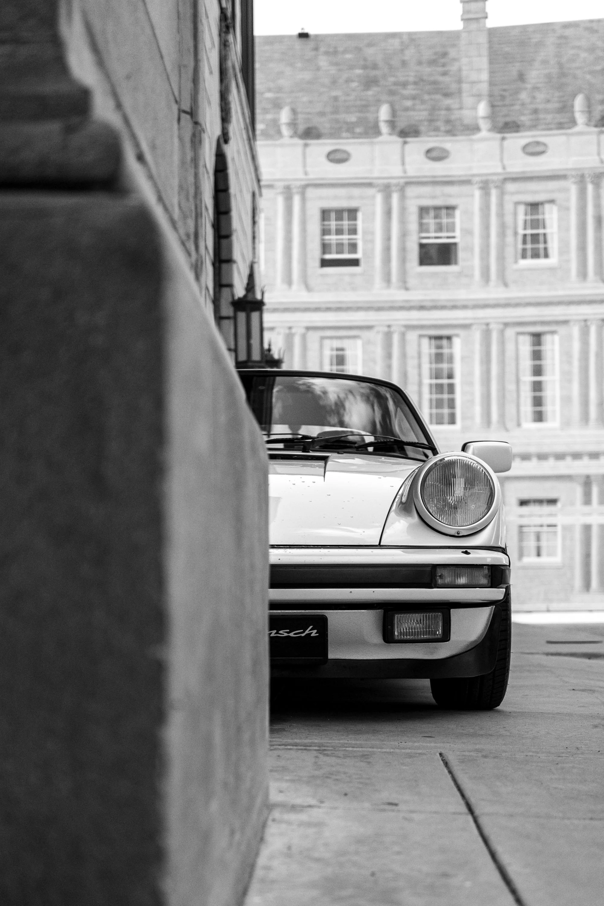 Black and white photo of an old Porsche car parked behind a low concrete wall, with a historic building in the background.