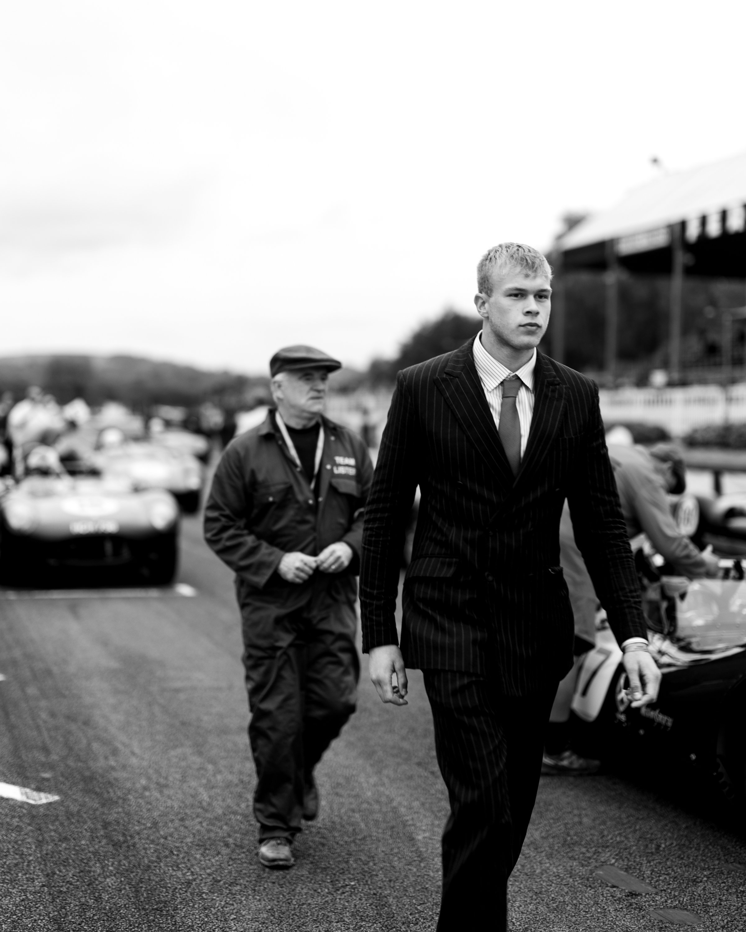 A black and white photo showing a young man with blond hair in a pinstripe suit and tie walking past an older man in casual clothing, with a background of cars and a building.