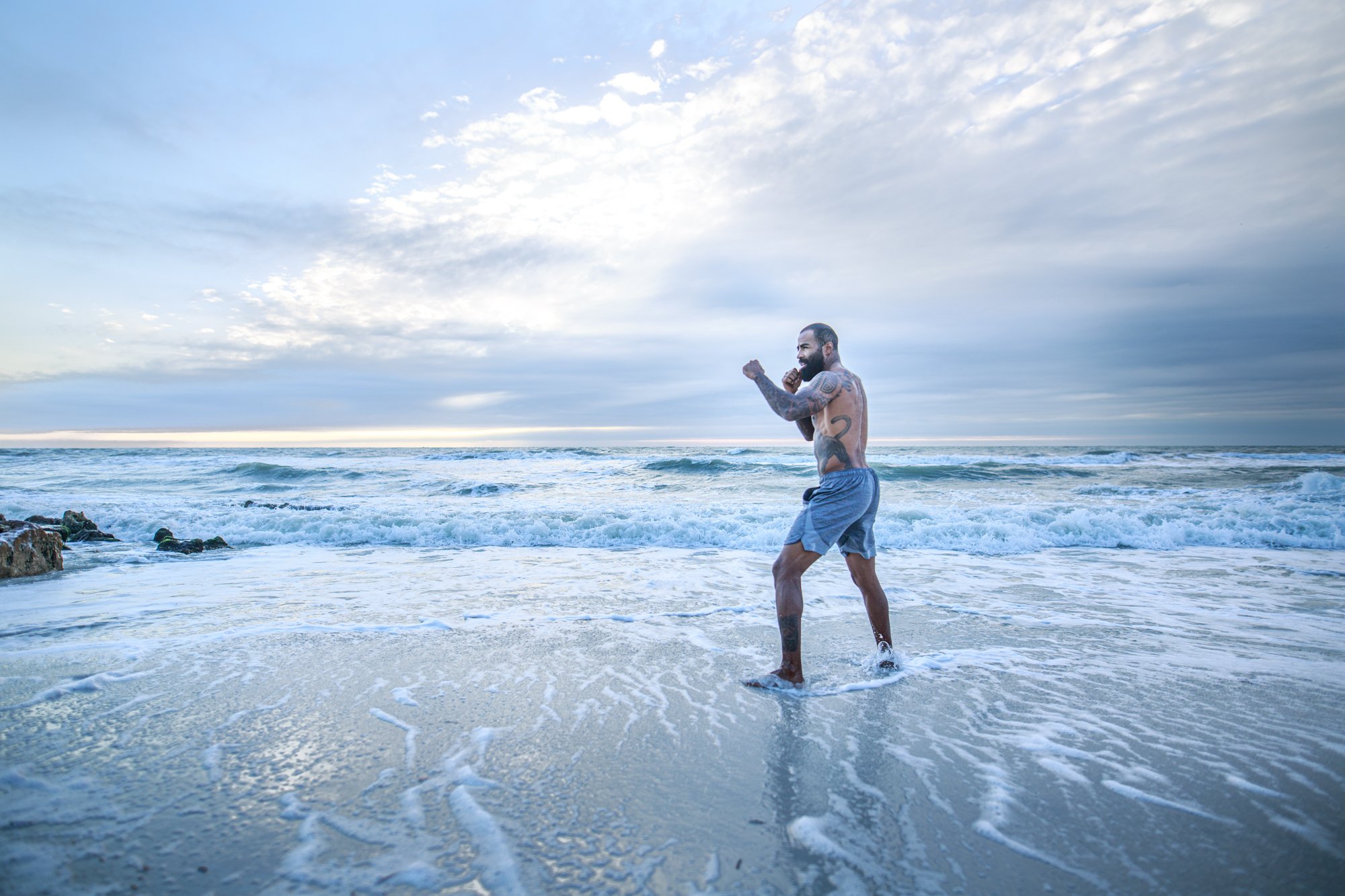 A muscular man with tattoos punching his fists in a boxing stance while standing in the ocean near the shore during sunset, with cloudy skies overhead.
