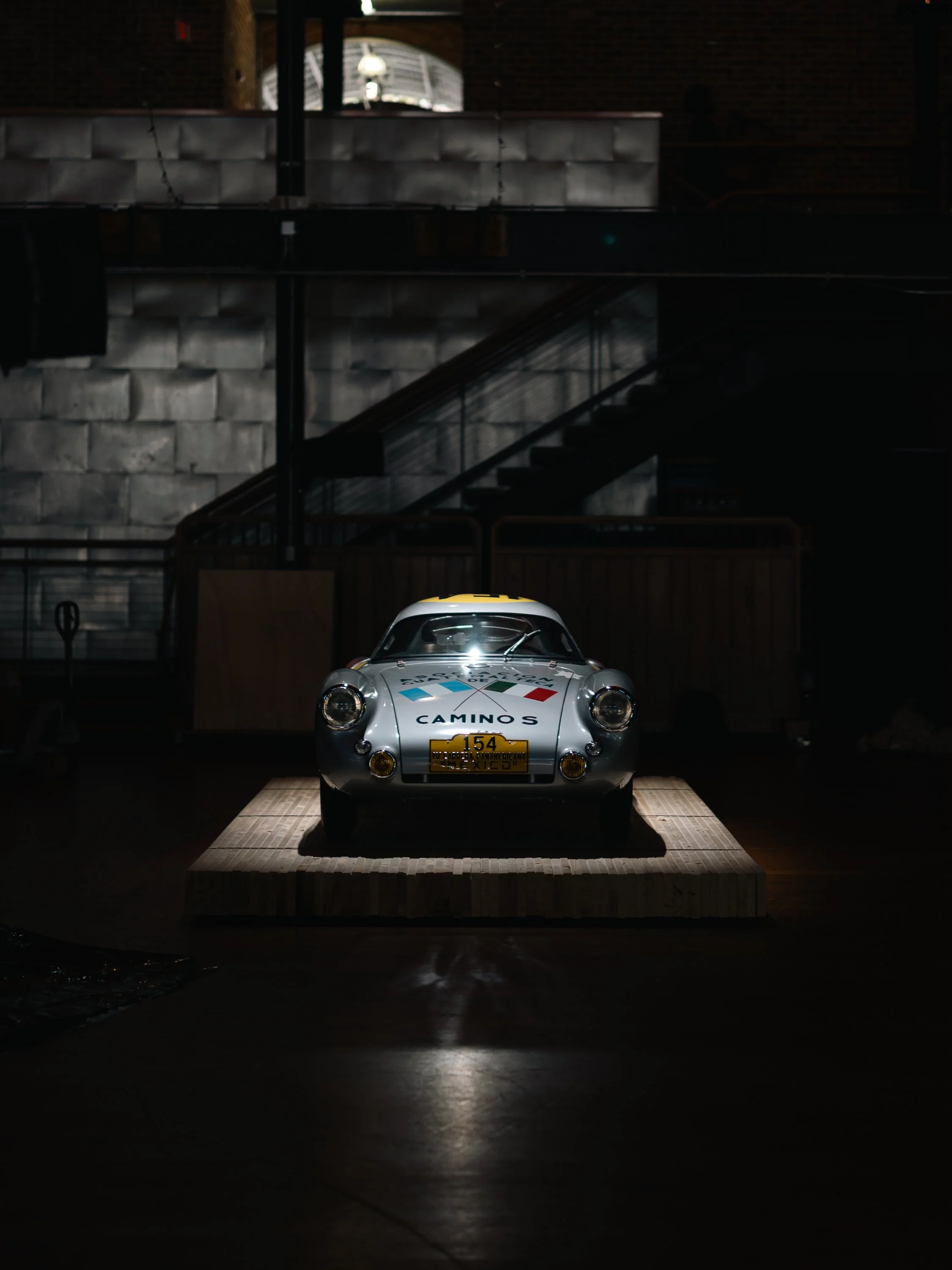 A vintage silver race car with a yellow roof, displaying the words 'Camino's' and the number 154, is illuminated and placed on a wooden platform inside a dark, industrial-style building. The car has a license plate with the word 'Mexico' and the imag