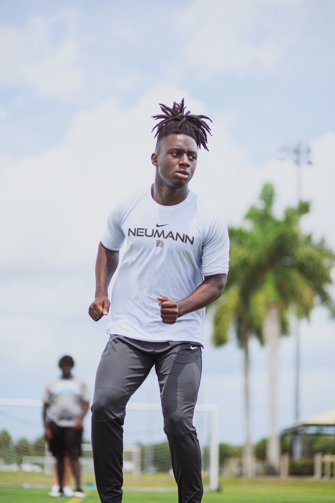 Young man in athletic gear, wearing a white NEUMANN shirt, standing on a sports field with trees and a fence in the background, appears to be running or exercising outdoors.