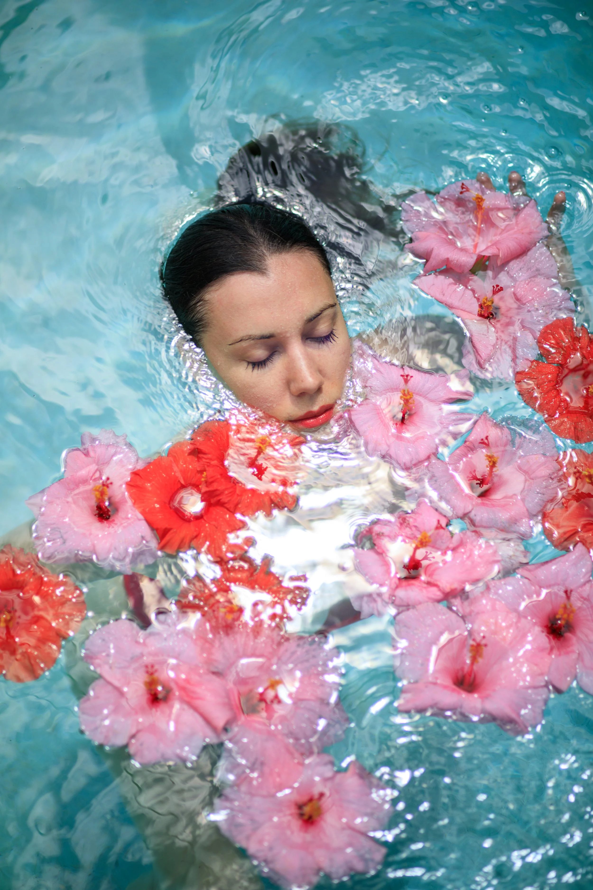 A woman with closed eyes floating in a swimming pool, surrounded by pink and red hibiscus flowers. Vogue italia.