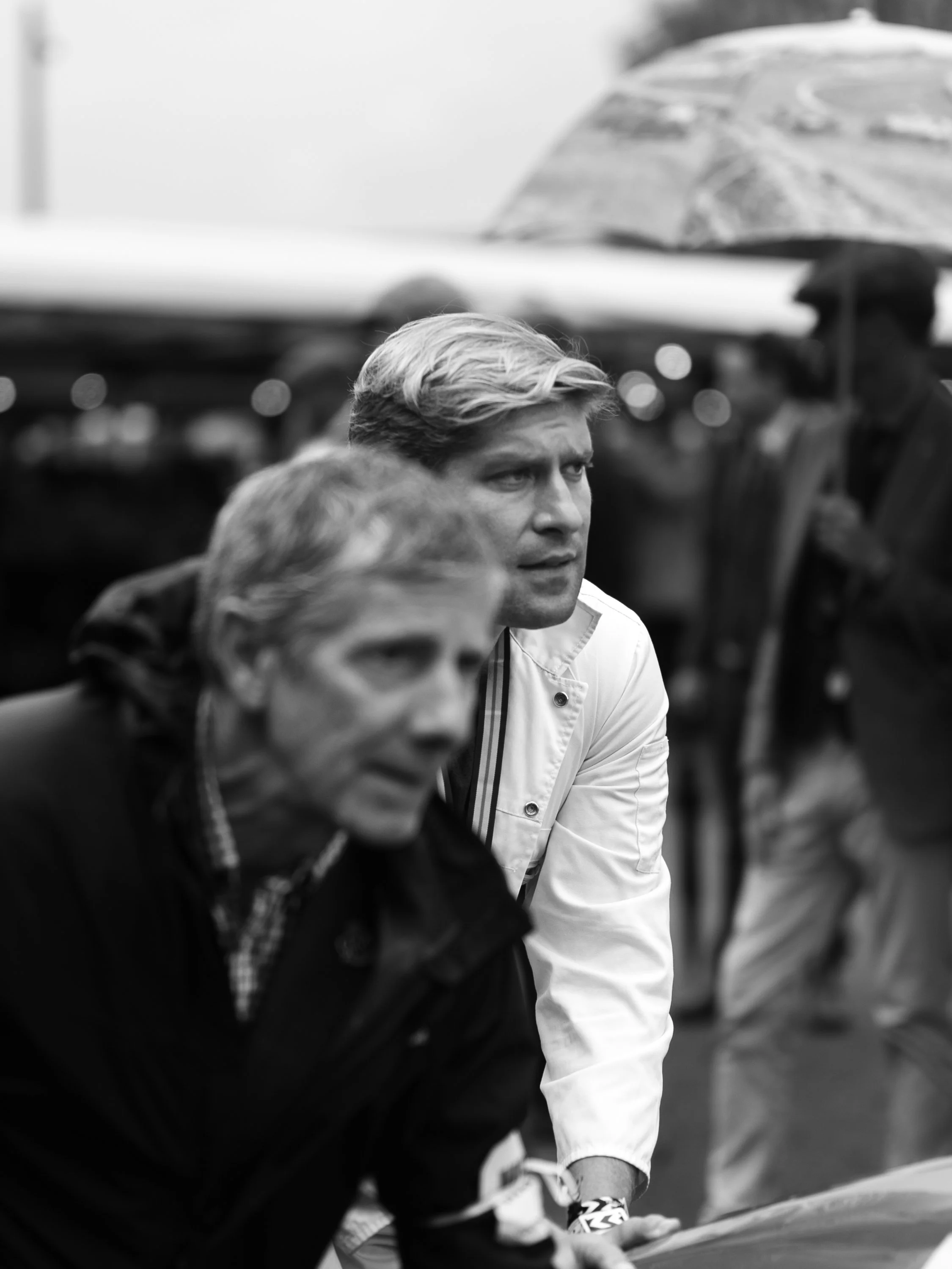 Black and white photo of two men, one in a white jacket, sitting and focused, with blurred background including a person holding an umbrella.