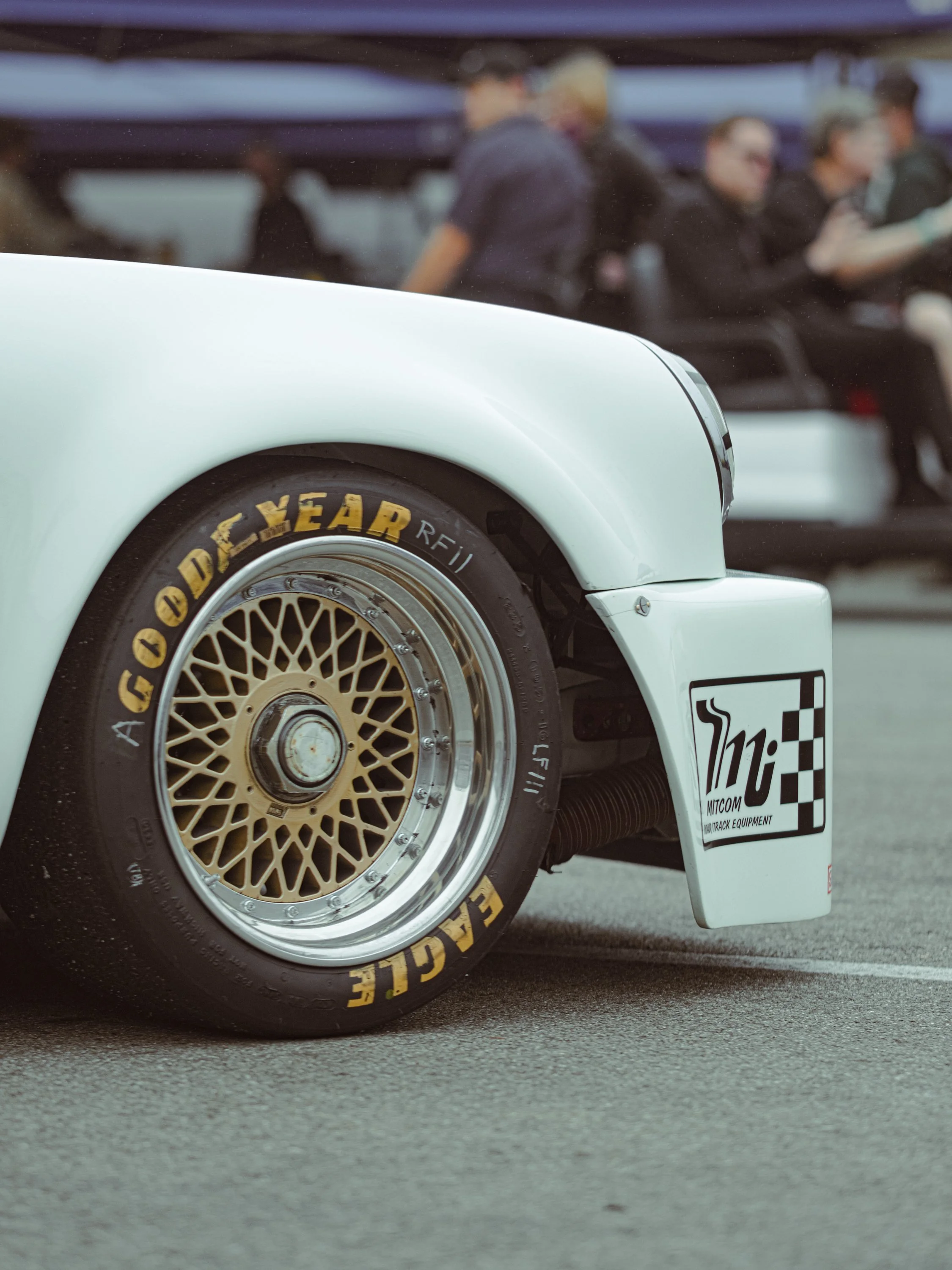 Close-up of a race car's front wheel with Goodyear branding and a detailed rim, set on a tarmac surface, with a blurred background of people.