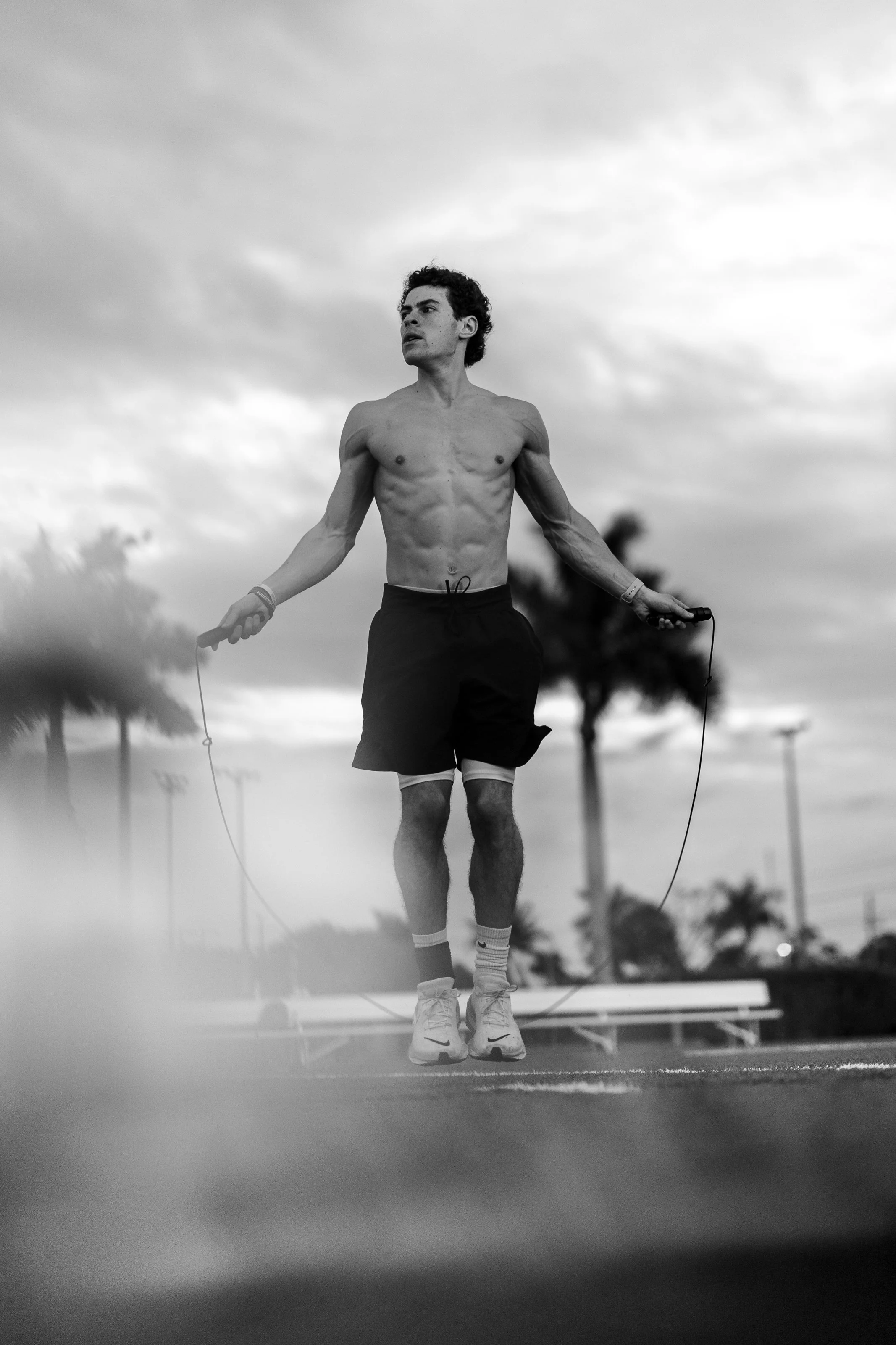 Black and white photo of a young man with curly hair skipping rope outdoors, wearing shorts, socks, and sneakers, with a cloudy sky and palm trees in the background.