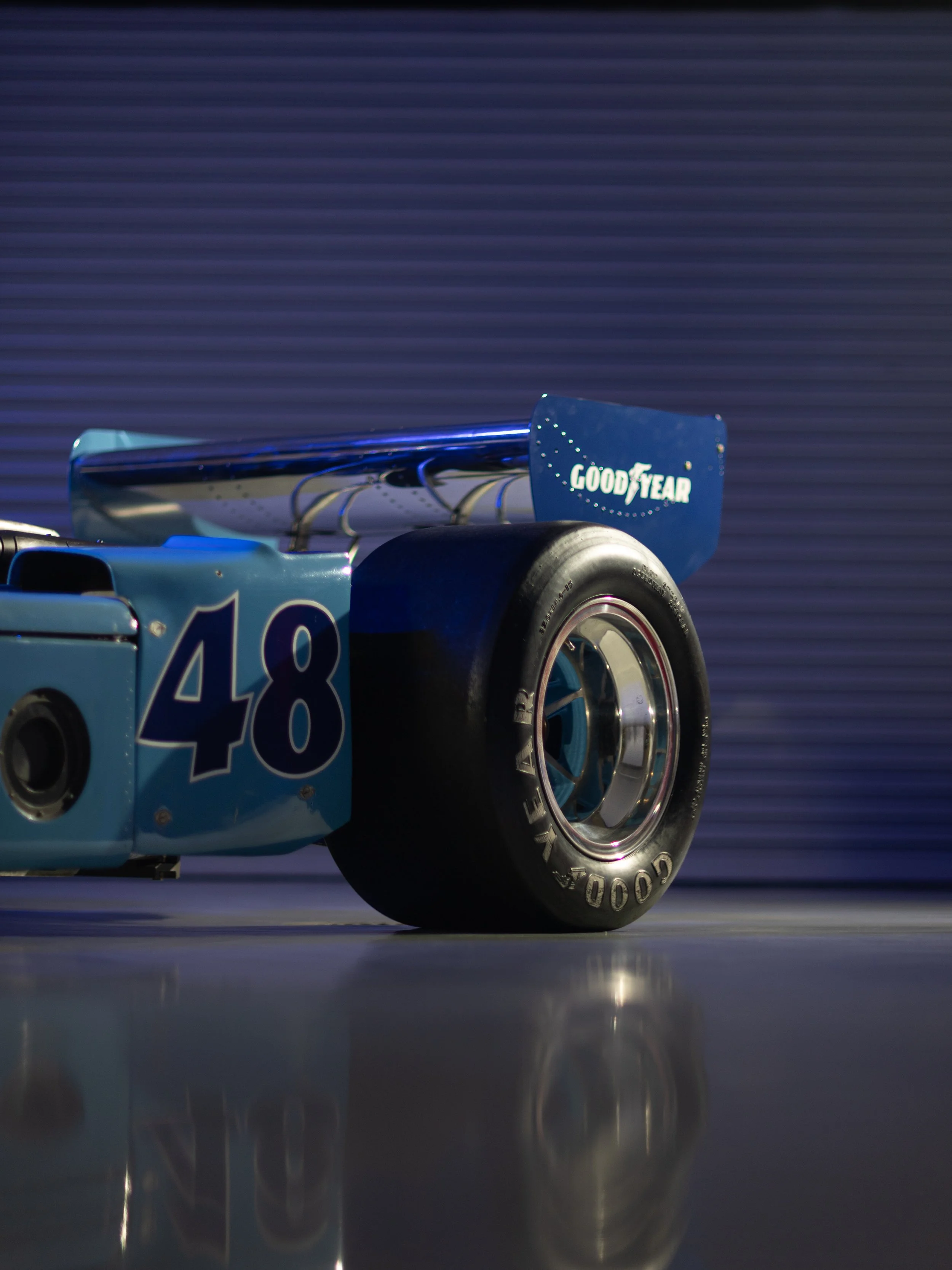 Close-up of a vintage race car with a prominent rear wing, tire branding, and the number 48 on the vehicle, on a reflective surface with a blurred background.