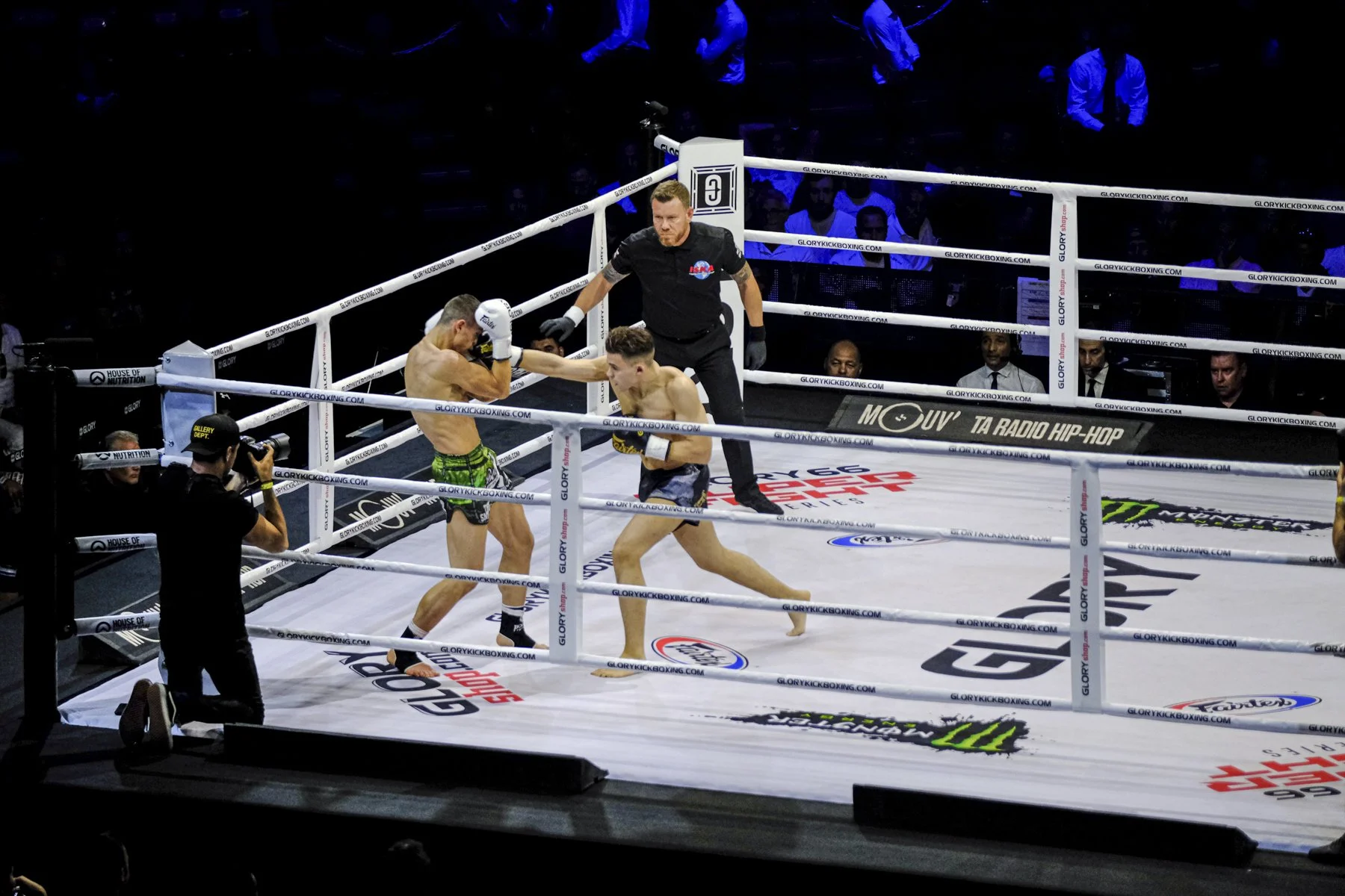 Two male fighters in a kickboxing match inside a boxing ring, one throwing a punch at the other. A referee stands nearby observing the action. Spectators are visible in the background, with some taking photos.