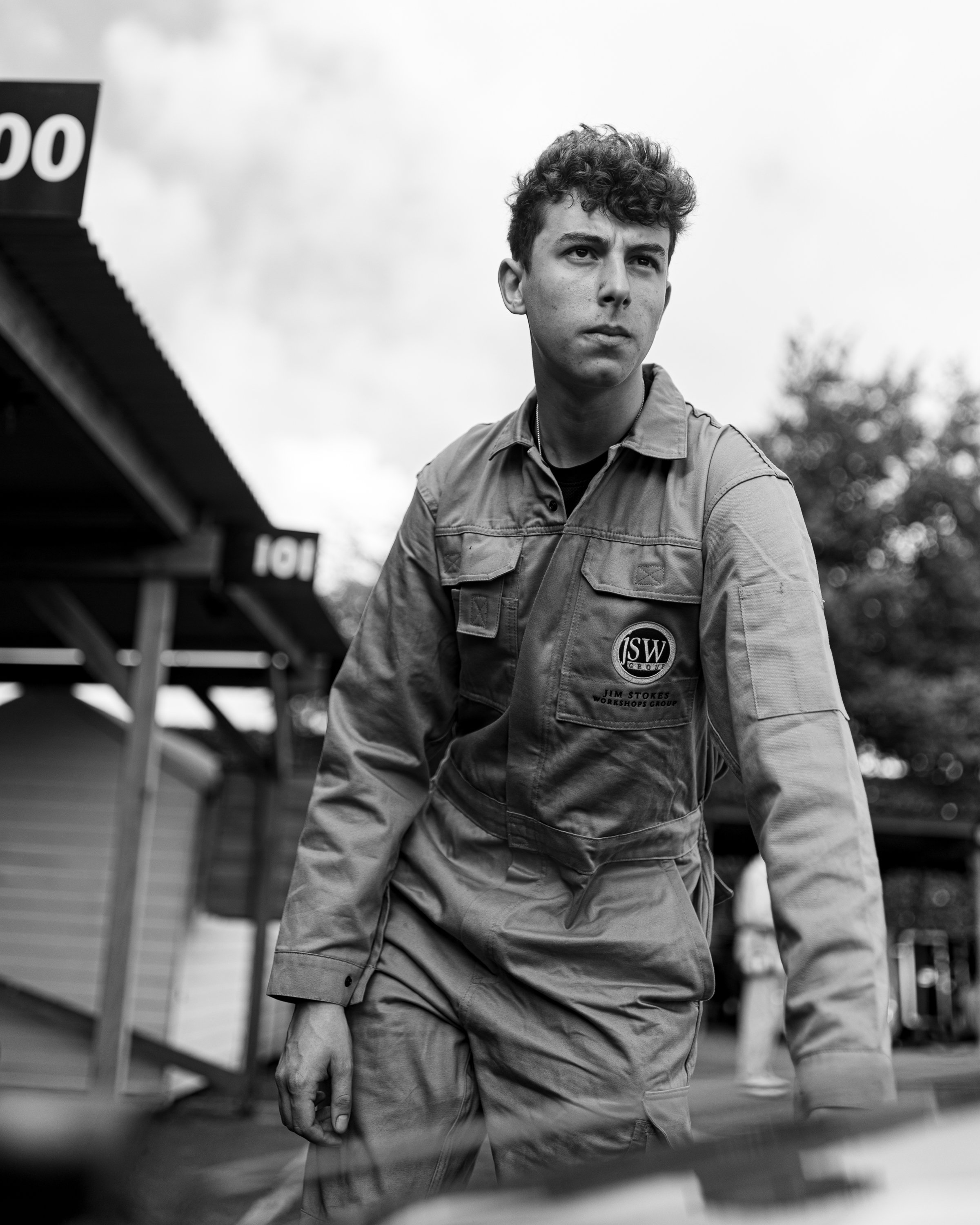 A young man with curly hair and a serious expression, wearing a jumpsuit with a patch that reads 'JSW Group' and 'Jim Stokes Workshops Group', standing outdoors with a cloudy sky and trees in the background.