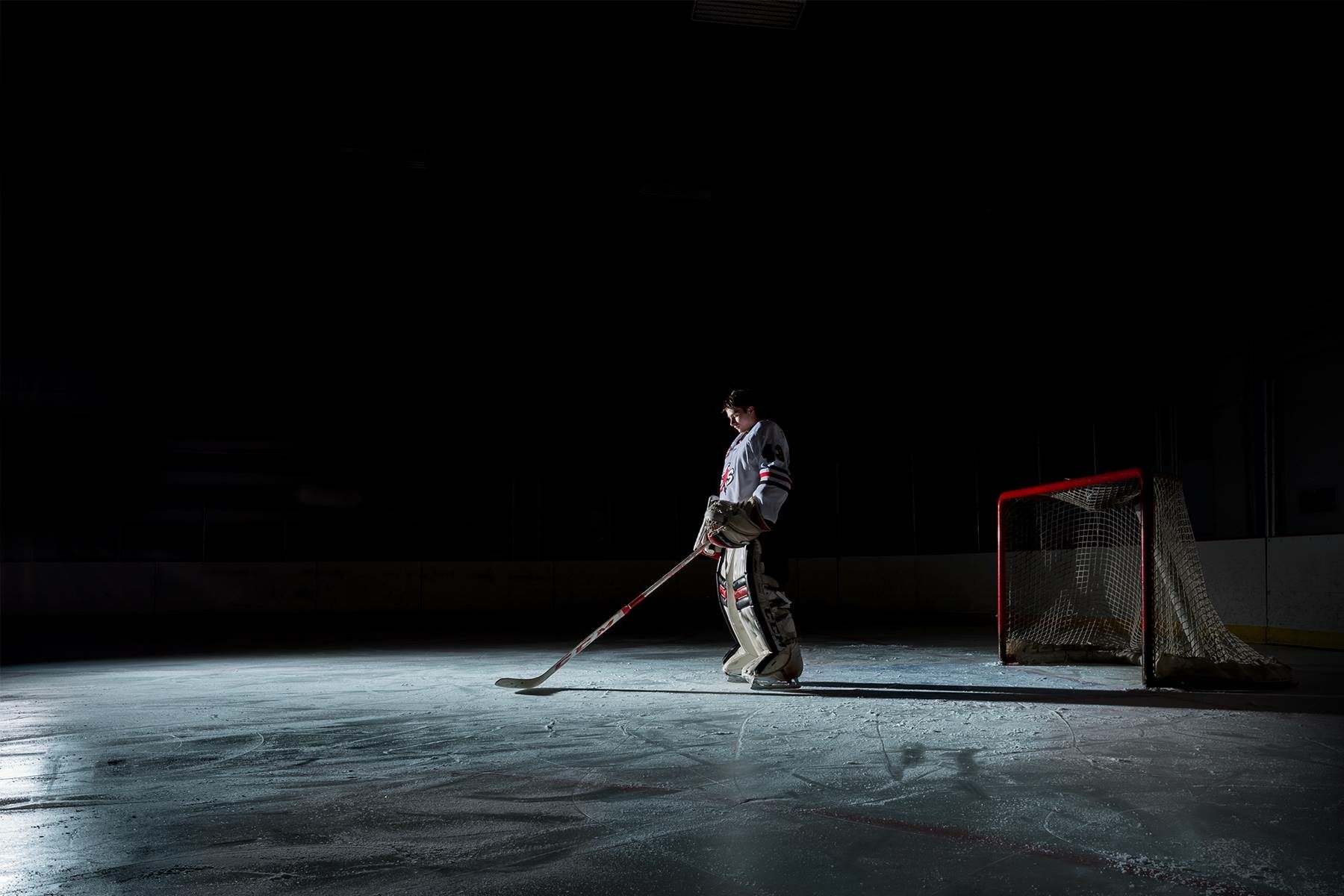 A hockey goalie standing alone on the ice rink behind the goal net, with dramatic lighting creating high contrast and shadows.