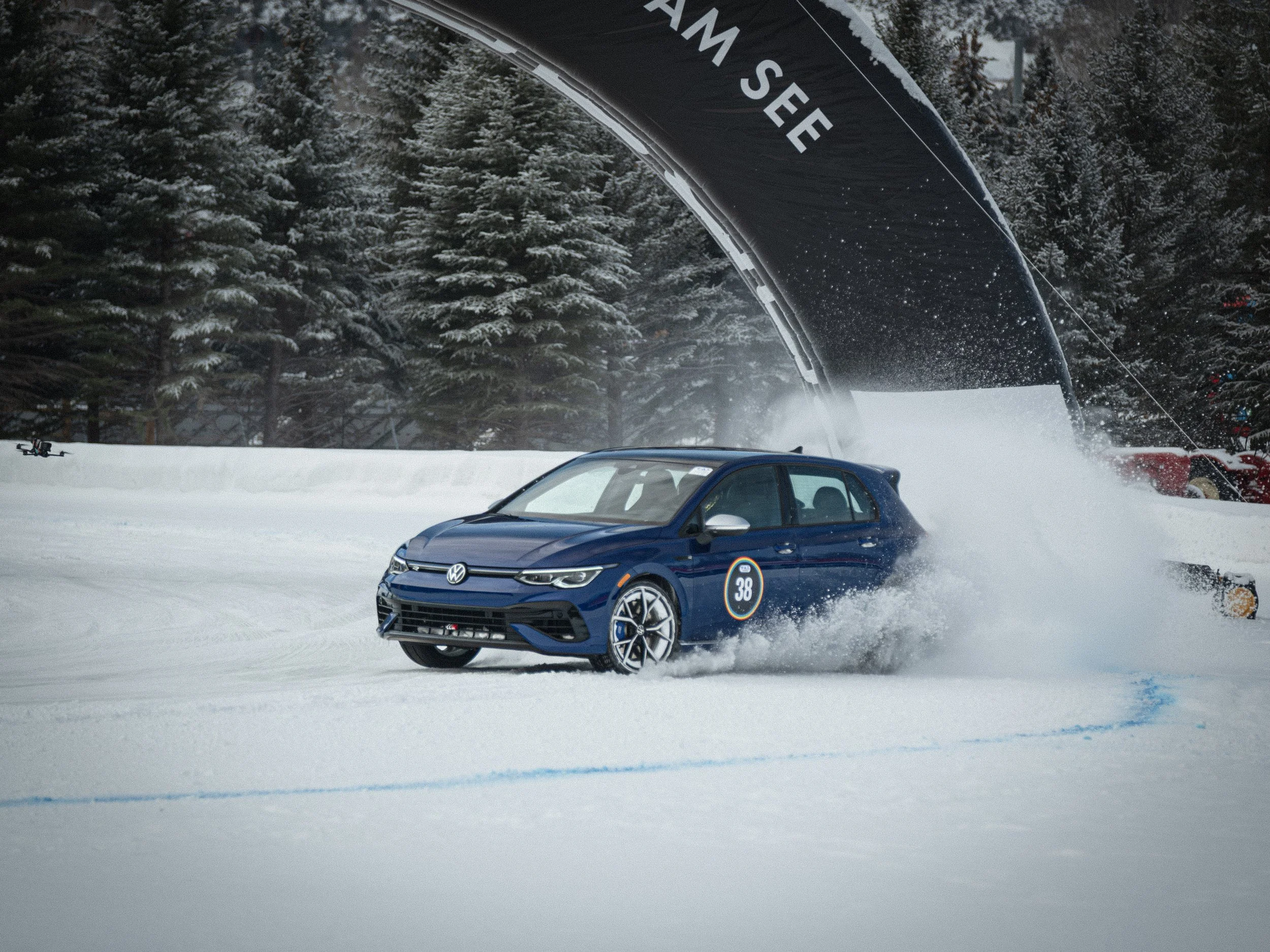 A blue Volkswagen sedan speeding on a snow-covered road in a snowy forest, with snow spray behind it, during winter daytime. VW Golf R driven by Tanner Foust, Fat Ice Race, Aspen Colorado