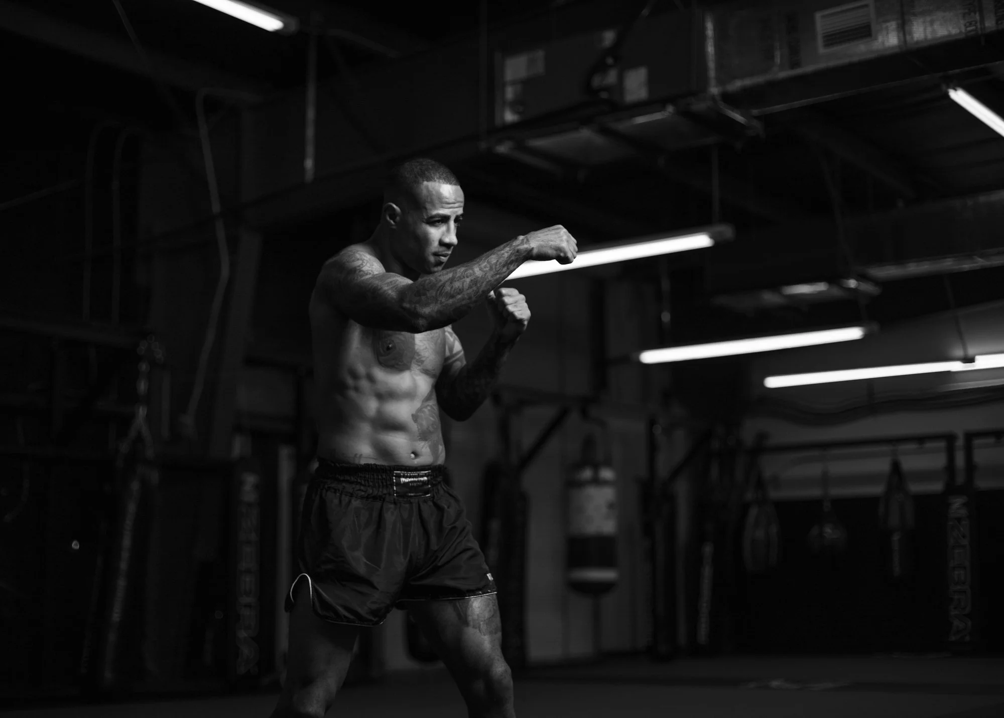 A shirtless man with tattoos on his arms and torso, wearing shorts, is boxing or practicing punches inside a gym with a dark industrial background, illuminated by overhead lights. Cosmo Alexandre, muay thai