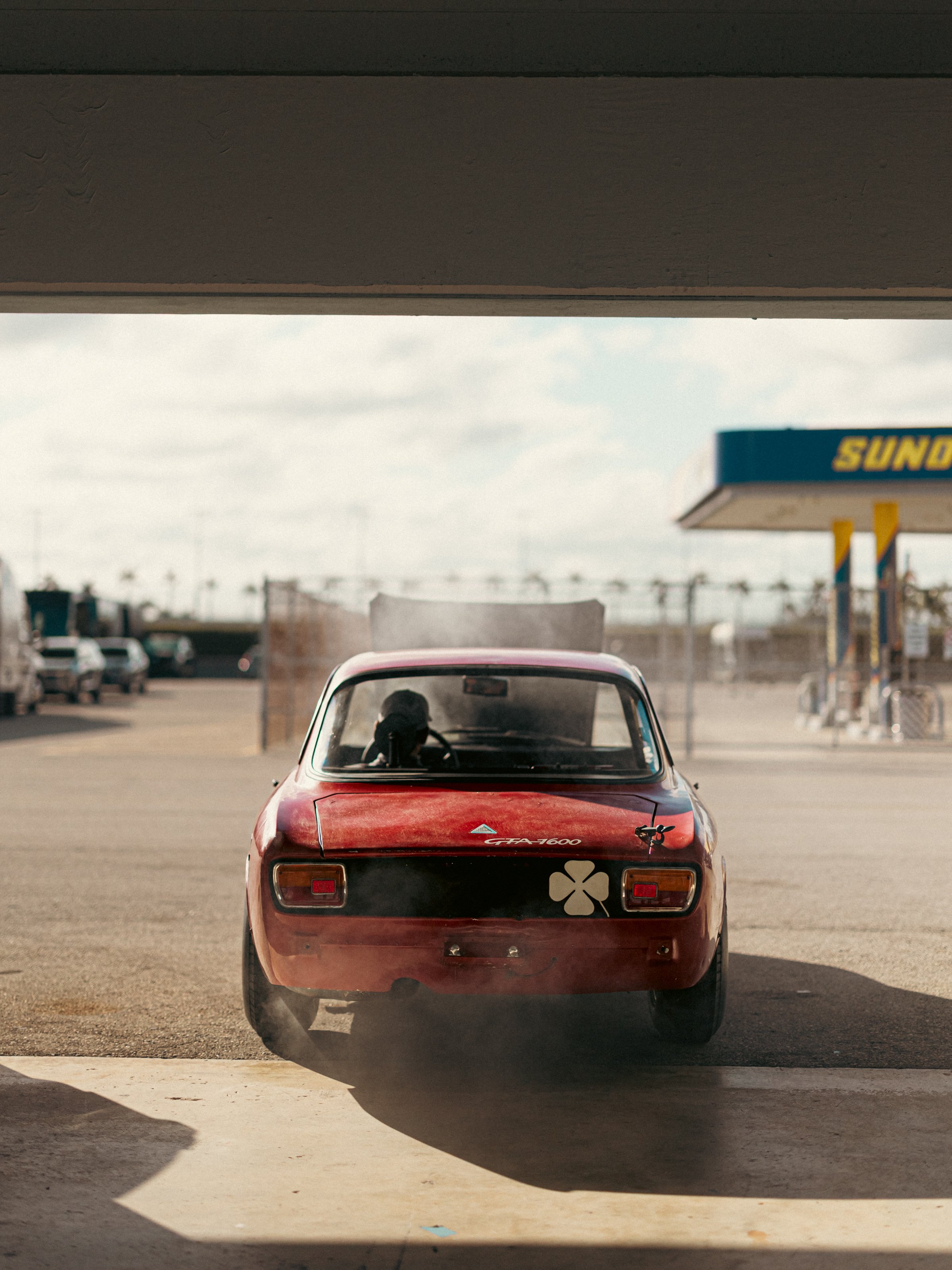 Red vintage car viewed from behind in a parking lot, with a large shadow cast over part of the scene and a store sign visible in the background.