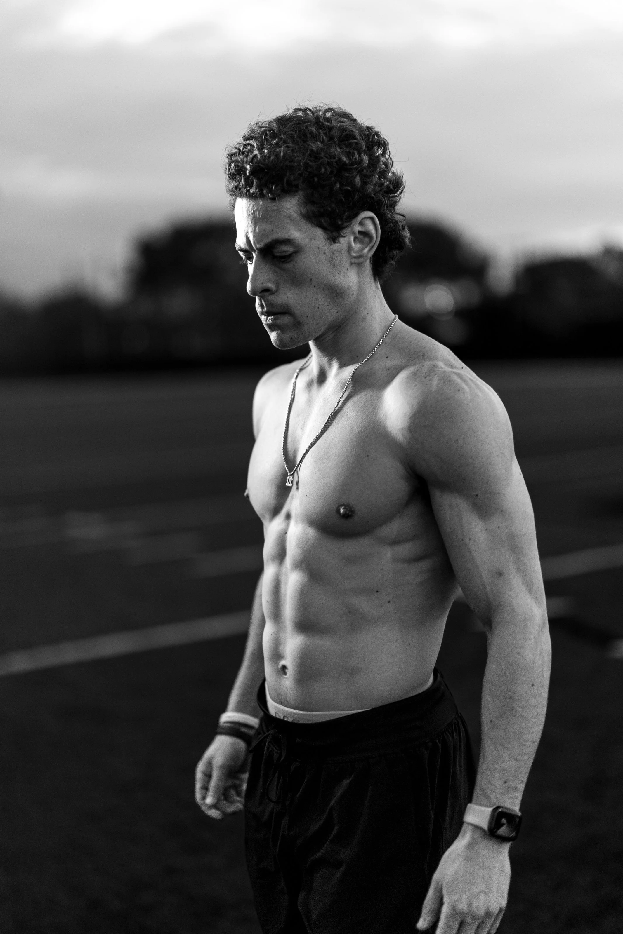 A young man with curly hair looking downward, shirtless, standing outdoors on a track field, wearing a watch and a necklace, with a blurred background of trees and sky.