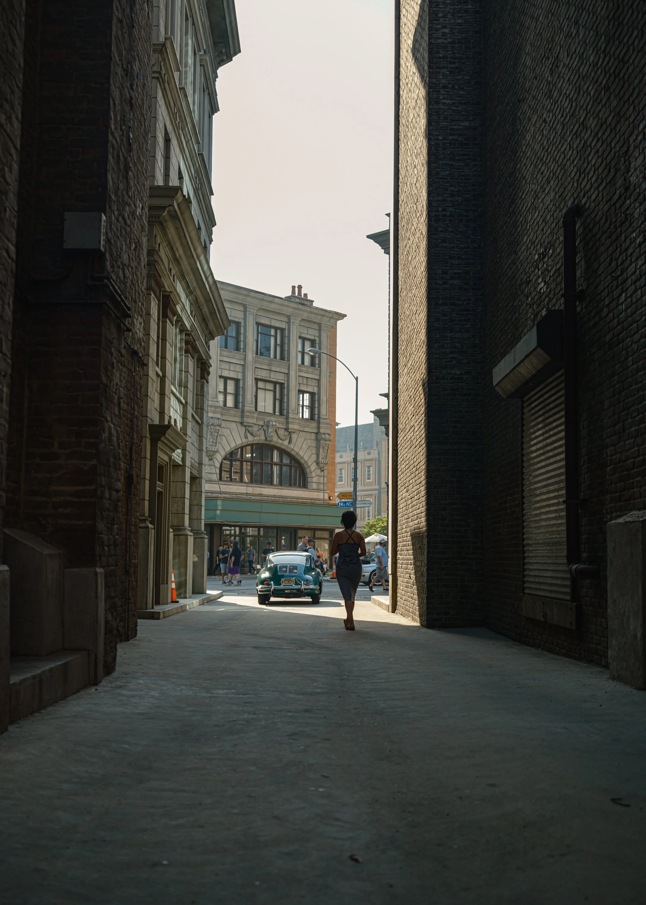 A narrow alleyway leading to a city street with a vintage green car parked in the foreground. In the background, a woman in a striped dress walks toward the street, and several pedestrians and a lamppost are visible under a partly cloudy sky.