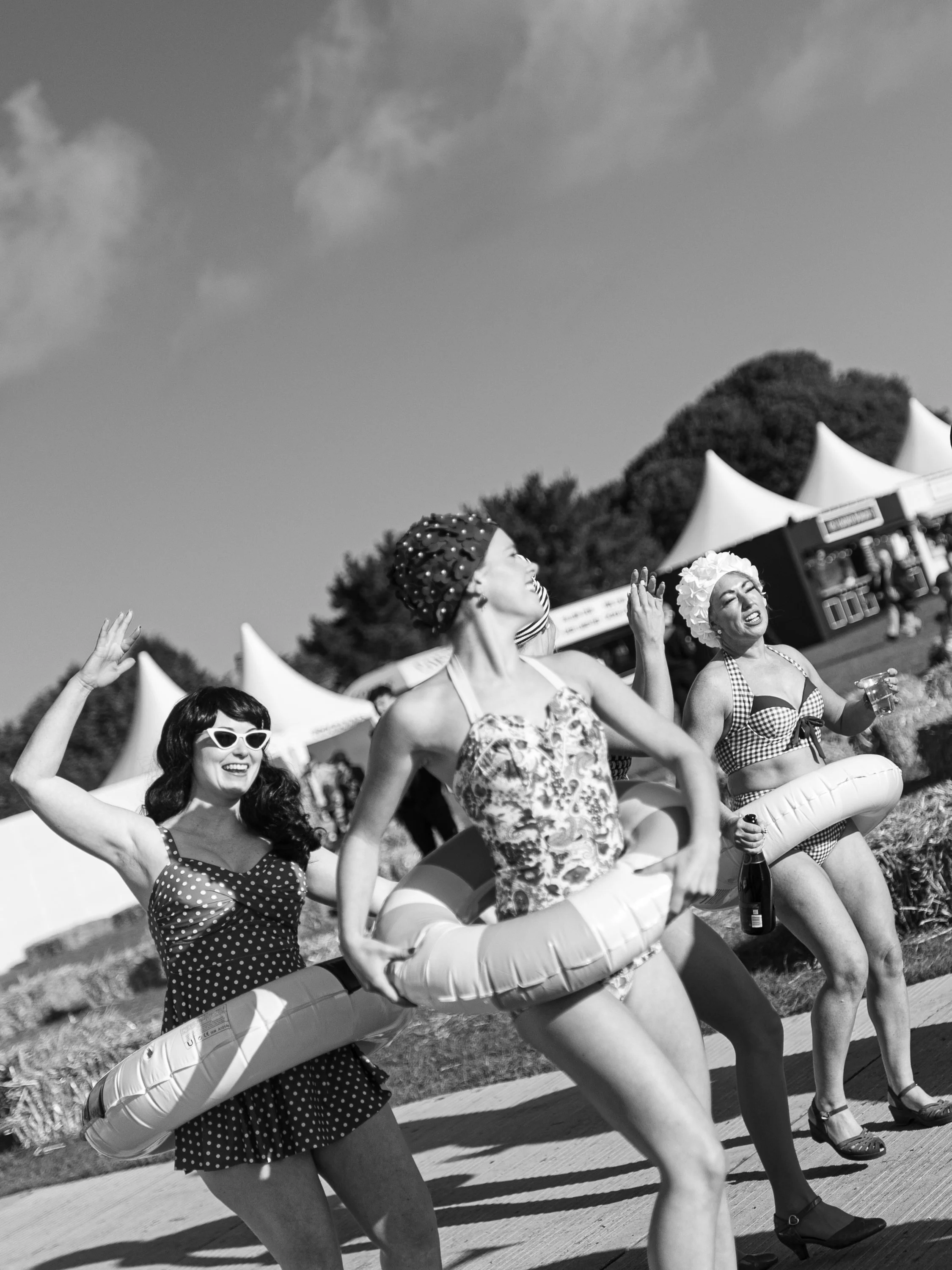 Women in vintage swimsuits, some with polka dots and checkered patterns, with inflatable swim rings, at an outdoor event with tents and trees in the background.