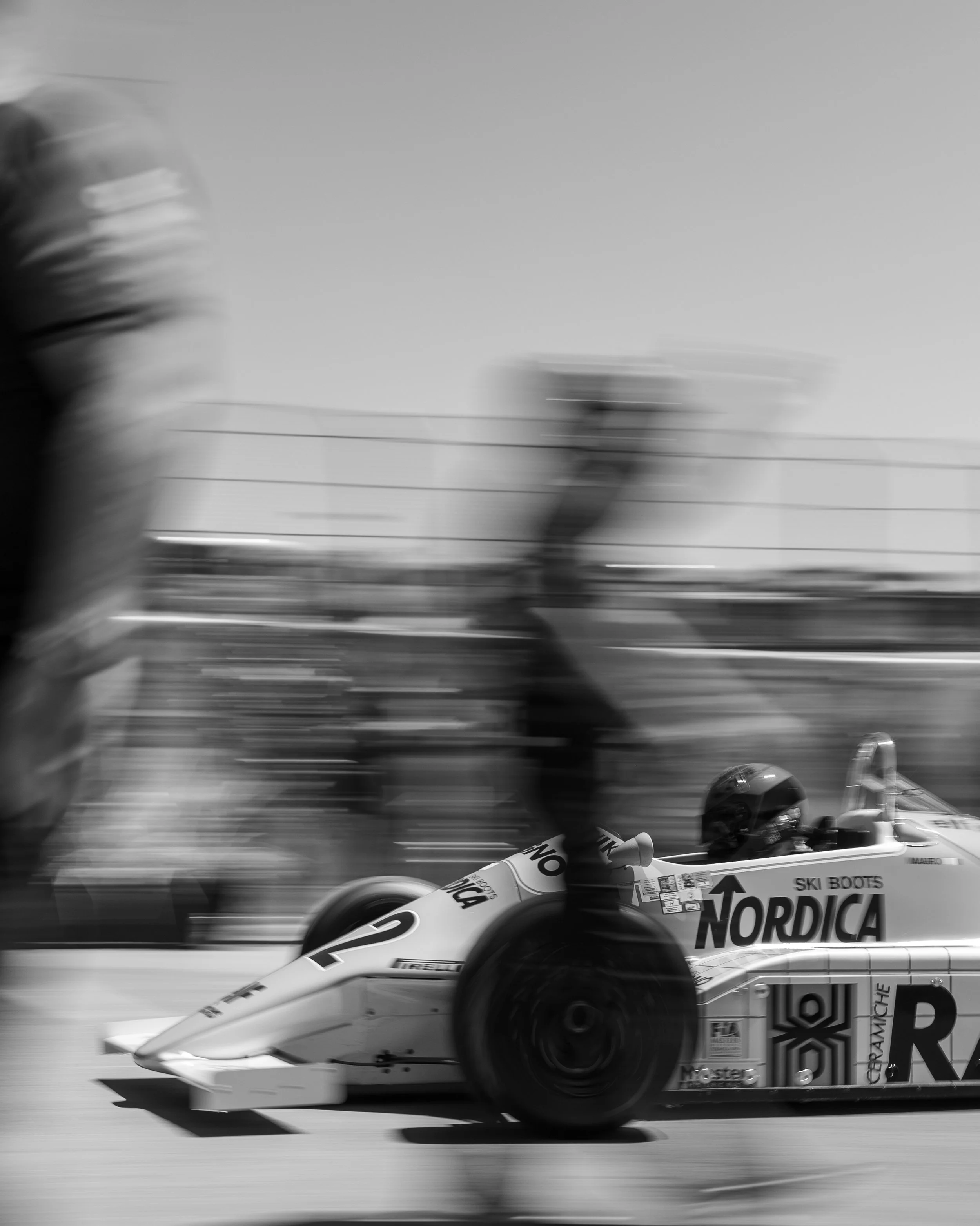 Black and white photo of a racing car on the track with sponsorship logos, blurred background indicating high speed.