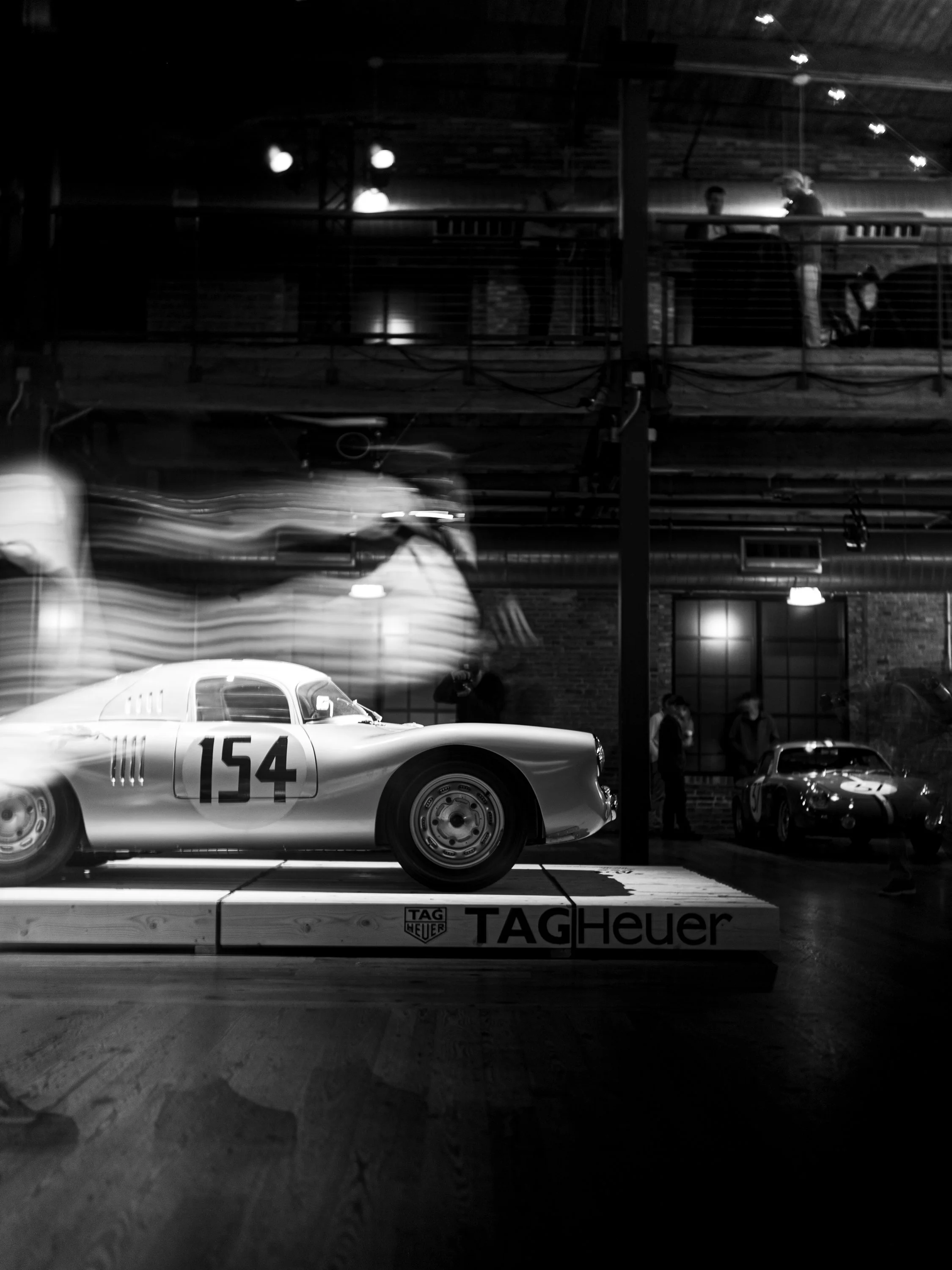 A vintage race car with the number 154 is displayed on a platform in a dimly lit indoor setting. There are people observing the car and a person taking a photo in the background. The platform has a TAG Heuer logo.