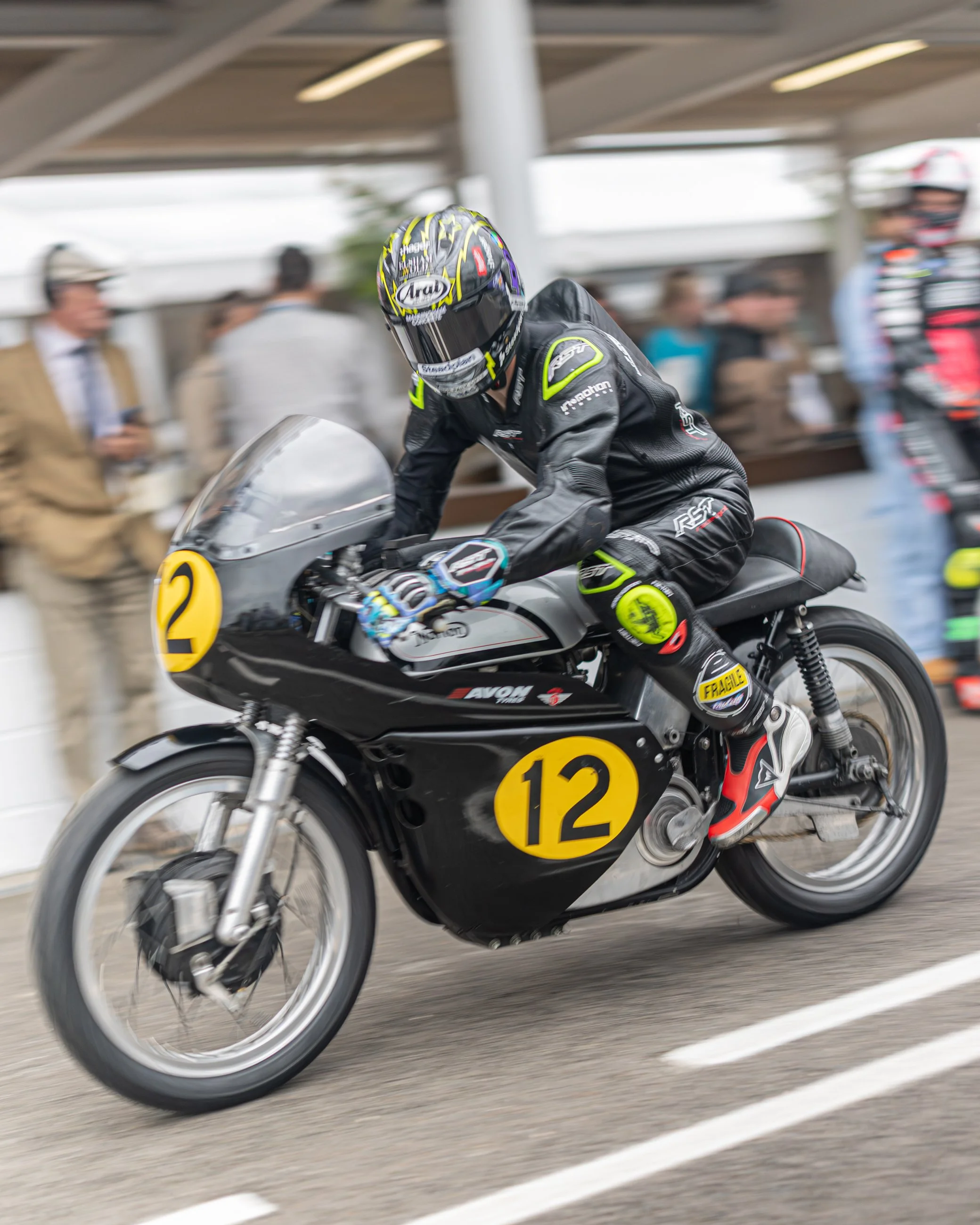 A motorcyclist in black racing gear and a colorful helmet riding a black motorcycle with yellow numbered decals, leaning forward on the motorcycle on a race track with blurred spectators and other riders in the background. Goodwood Revival