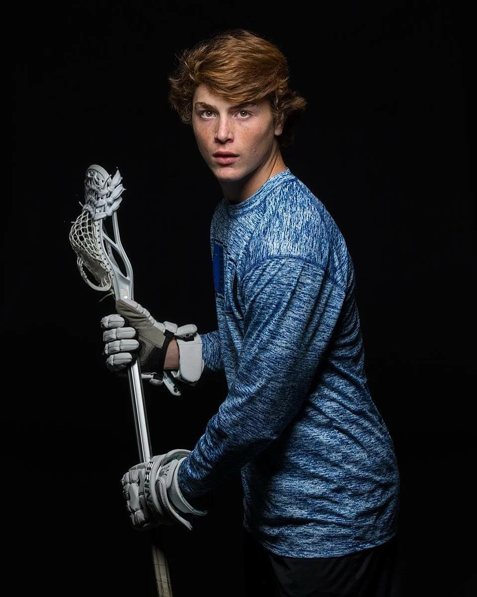 Young male lacrosse player holding a lacrosse stick, wearing gloves, with a serious expression, against a black background.