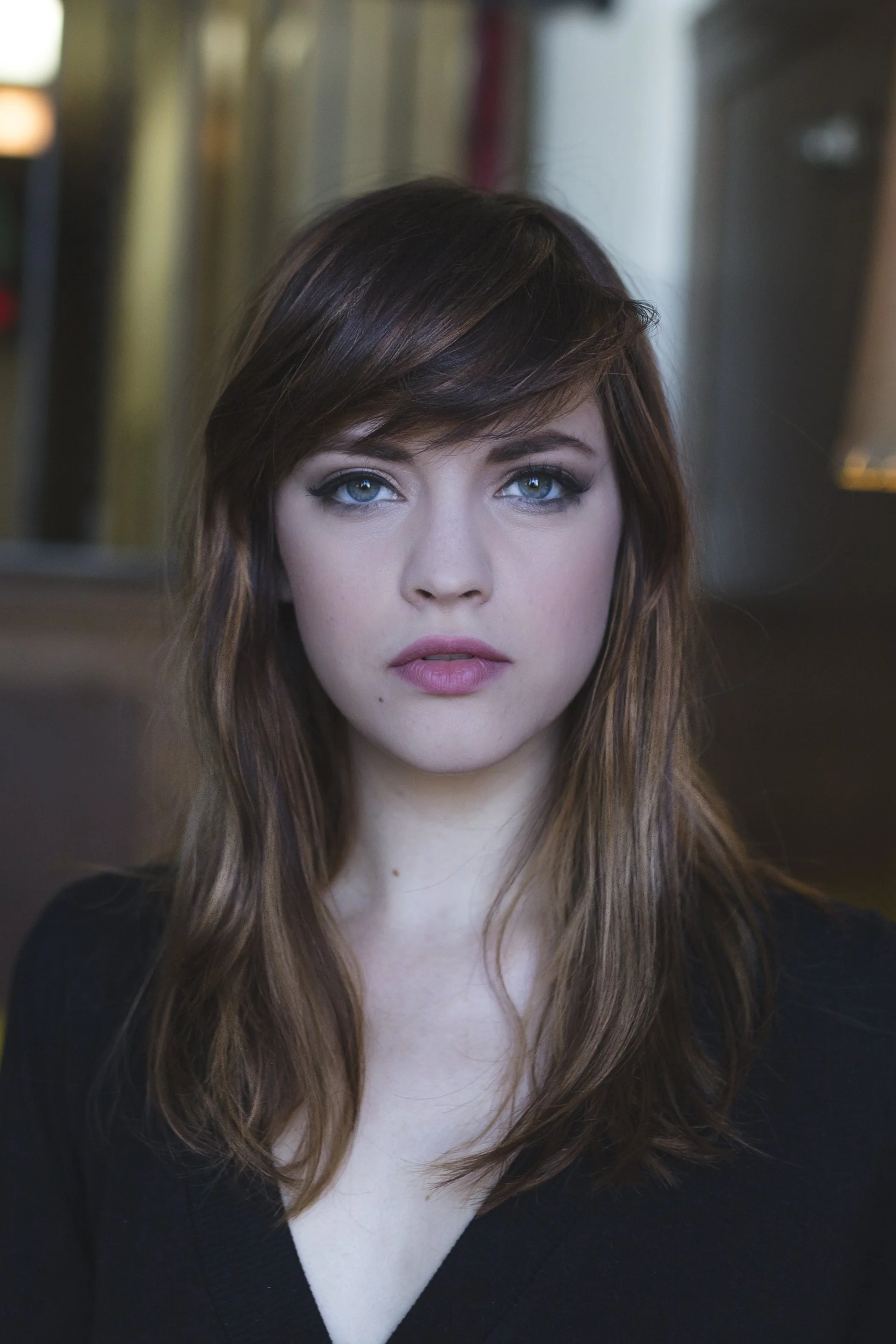 Portrait of a woman with blue eyes and long, wavy, reddish-brown hair, wearing a black top, with a blurred indoor background.