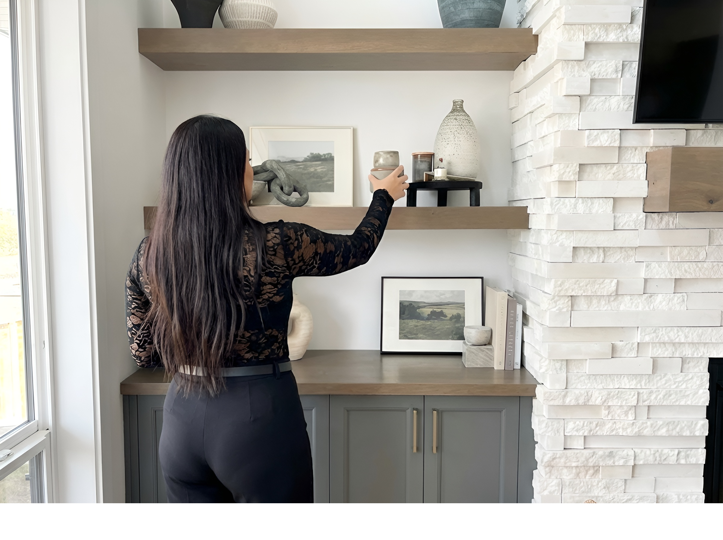 A woman with long dark hair, dressed in a black and brown patterned top and black pants, is facing a modern interior wall with decorative items on floating shelves, including vases and framed artwork.