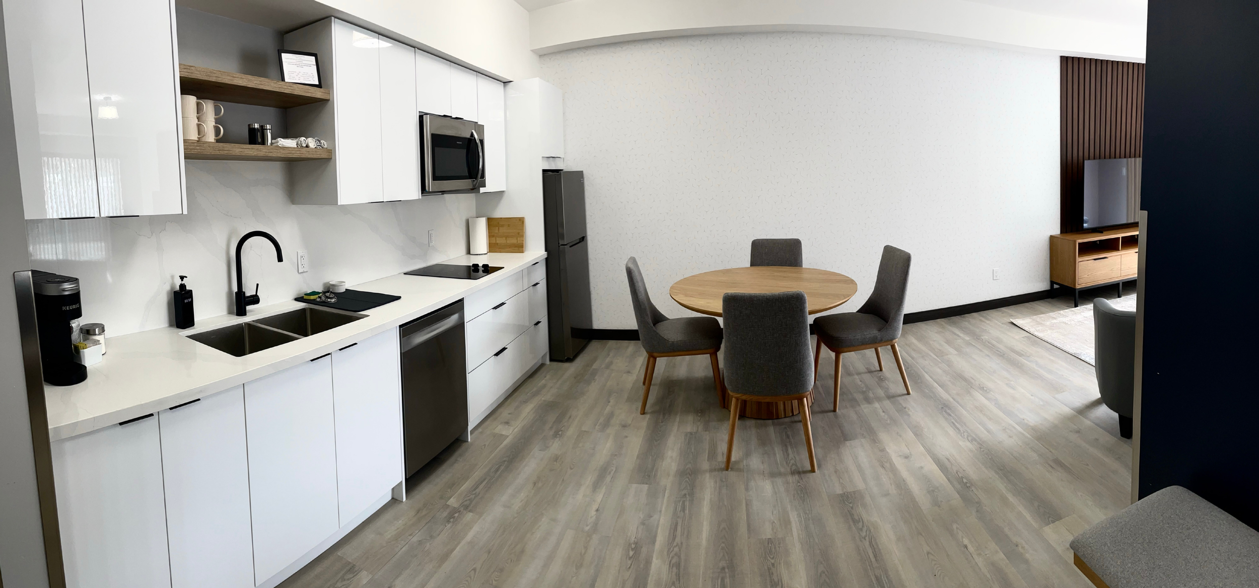 Modern kitchen with white cabinets, black sink, microwave, refrigerator, and open shelving; adjacent dining area with round wooden table and four gray upholstered chairs; living room with TV on wooden stand and seating area in the background.