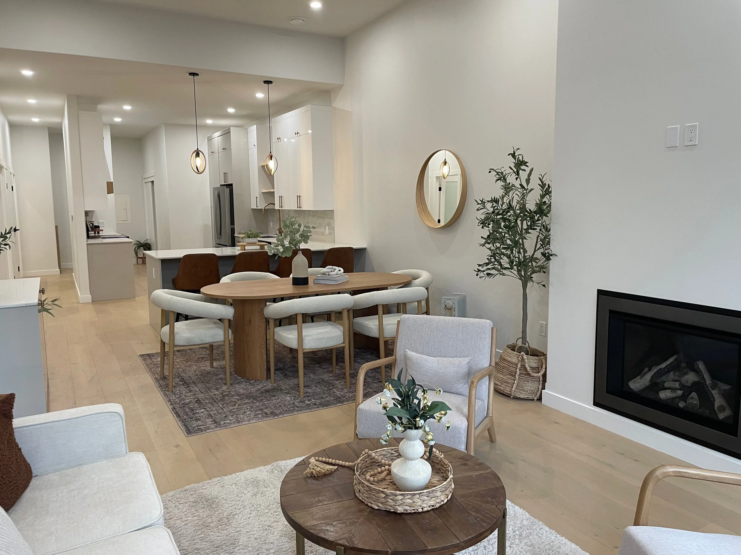 Open-concept living and dining area with white walls, light wood furniture, modern lighting fixtures, a fireplace, potted plants, and decorative accents.